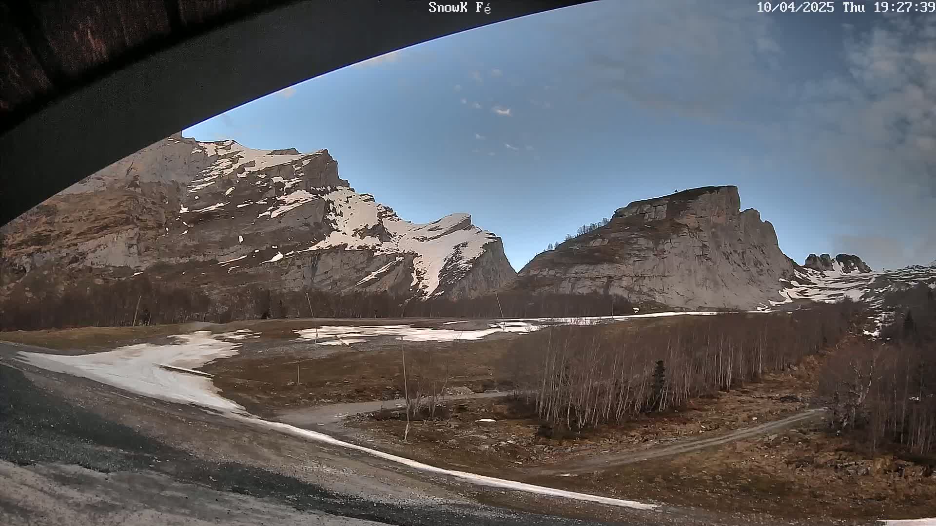 A mostly clear sky is over a valley with snow-dusted, rocky mountains and a sparsely wooded area with patches of snow on the ground.