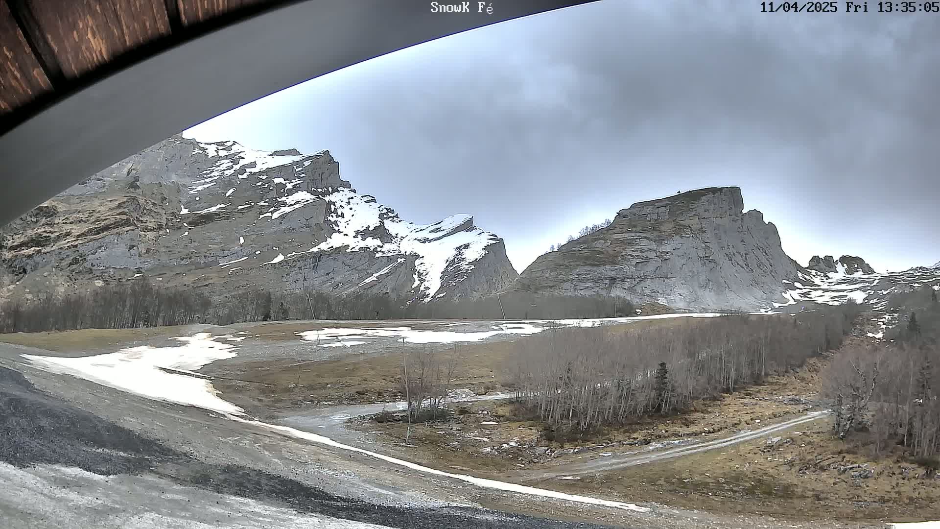 A partly cloudy day reveals a valley between two snow-dusted, rocky mountains with patches of snow on the ground and leafless trees.