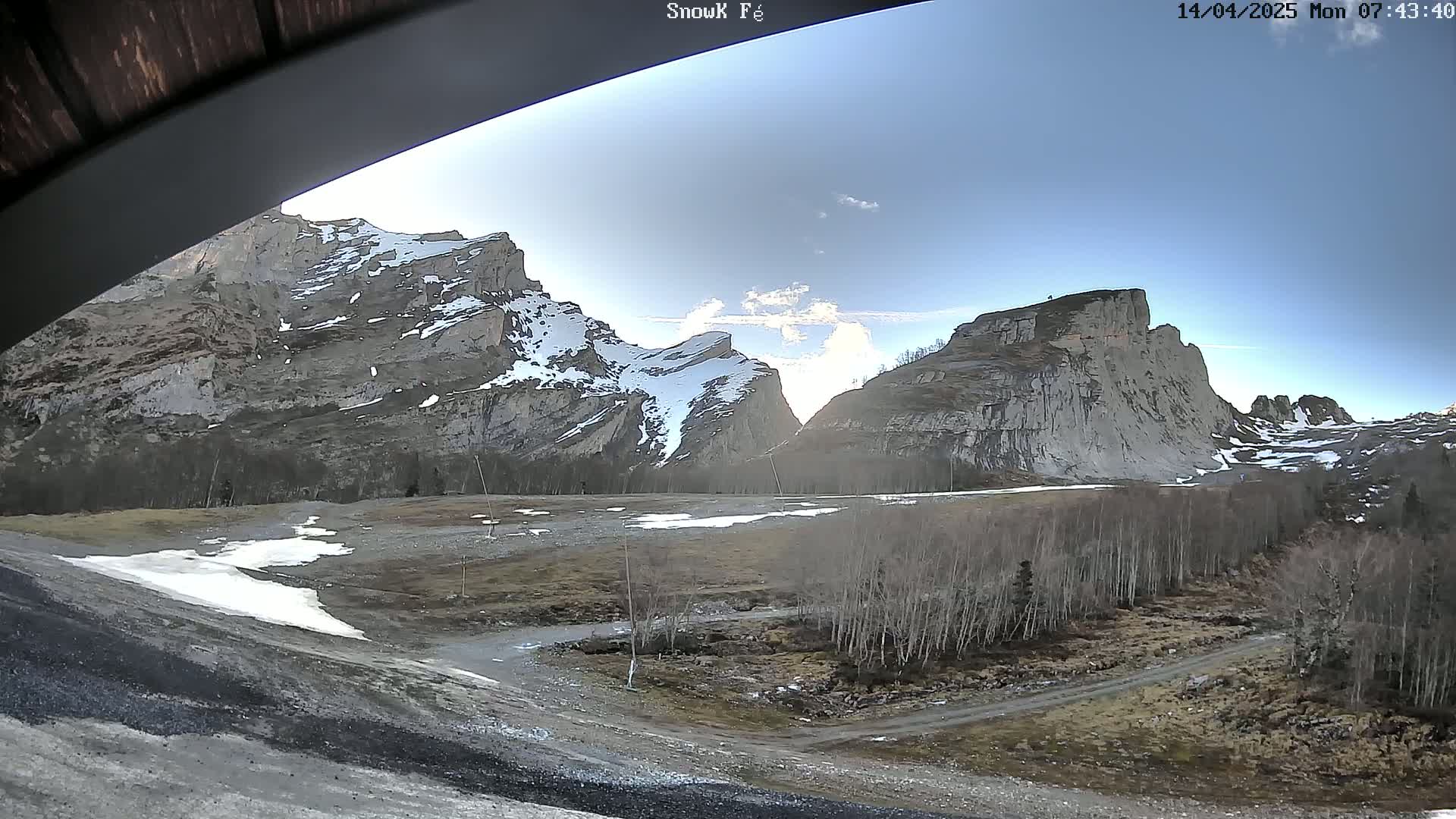 A mostly clear, sunny day reveals a valley floor with patchy snow and leafless trees, nestled between towering, snow-dusted mountain cliffs.