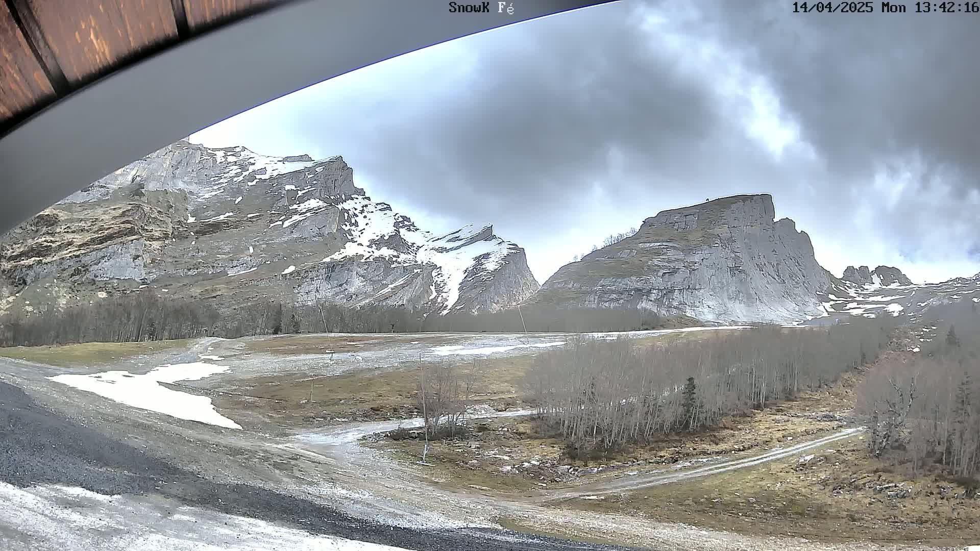 A view from under a covered structure shows snow-dusted, grey-colored mountains under a cloudy sky, with a flat, dirt area in the foreground containing patches of snow and sparse, leafless trees.