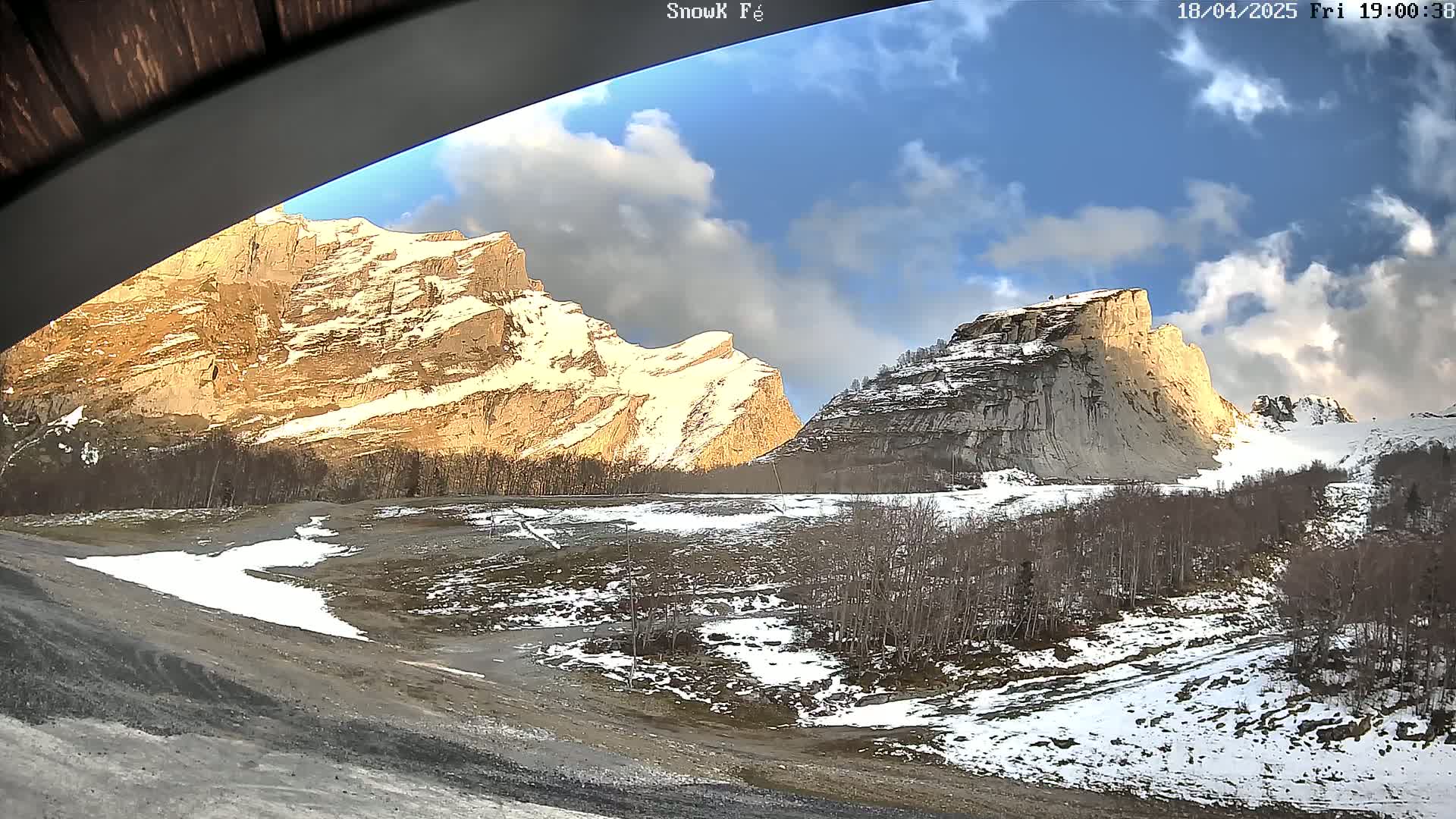 A partly cloudy sky illuminates snow-dusted mountains and a valley with patches of snow and bare trees.