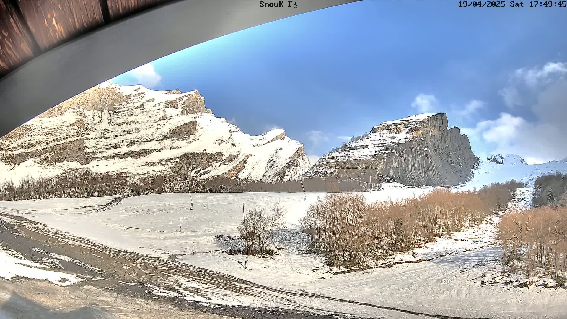 Snow-covered mountains and a valley with leafless trees under a mostly sunny sky.