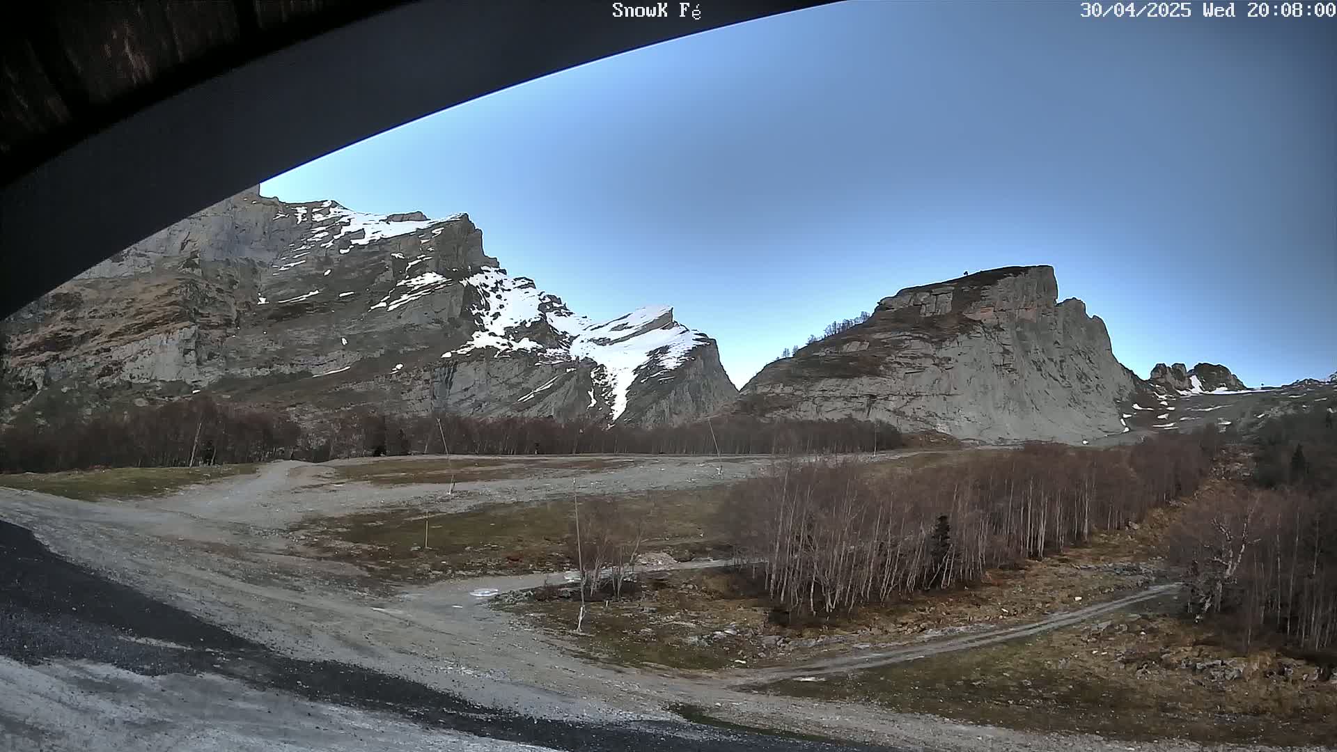 A mostly clear, blue sky overlooks a valley with a dirt road, leafless trees, and snow-capped rocky mountains in the background.