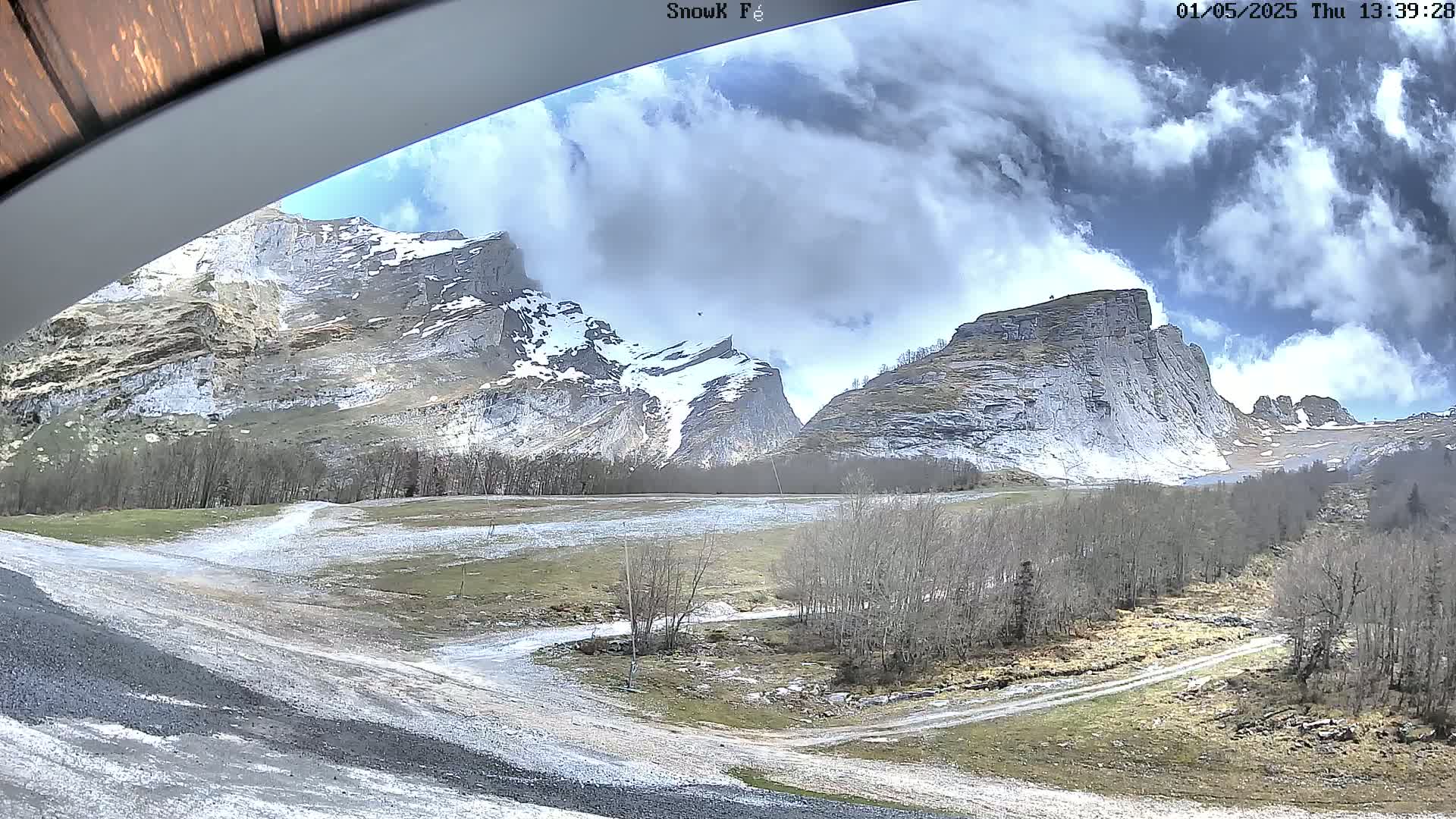 A partly cloudy day reveals snow-dusted mountains and a valley with a dirt road winding through bare trees.