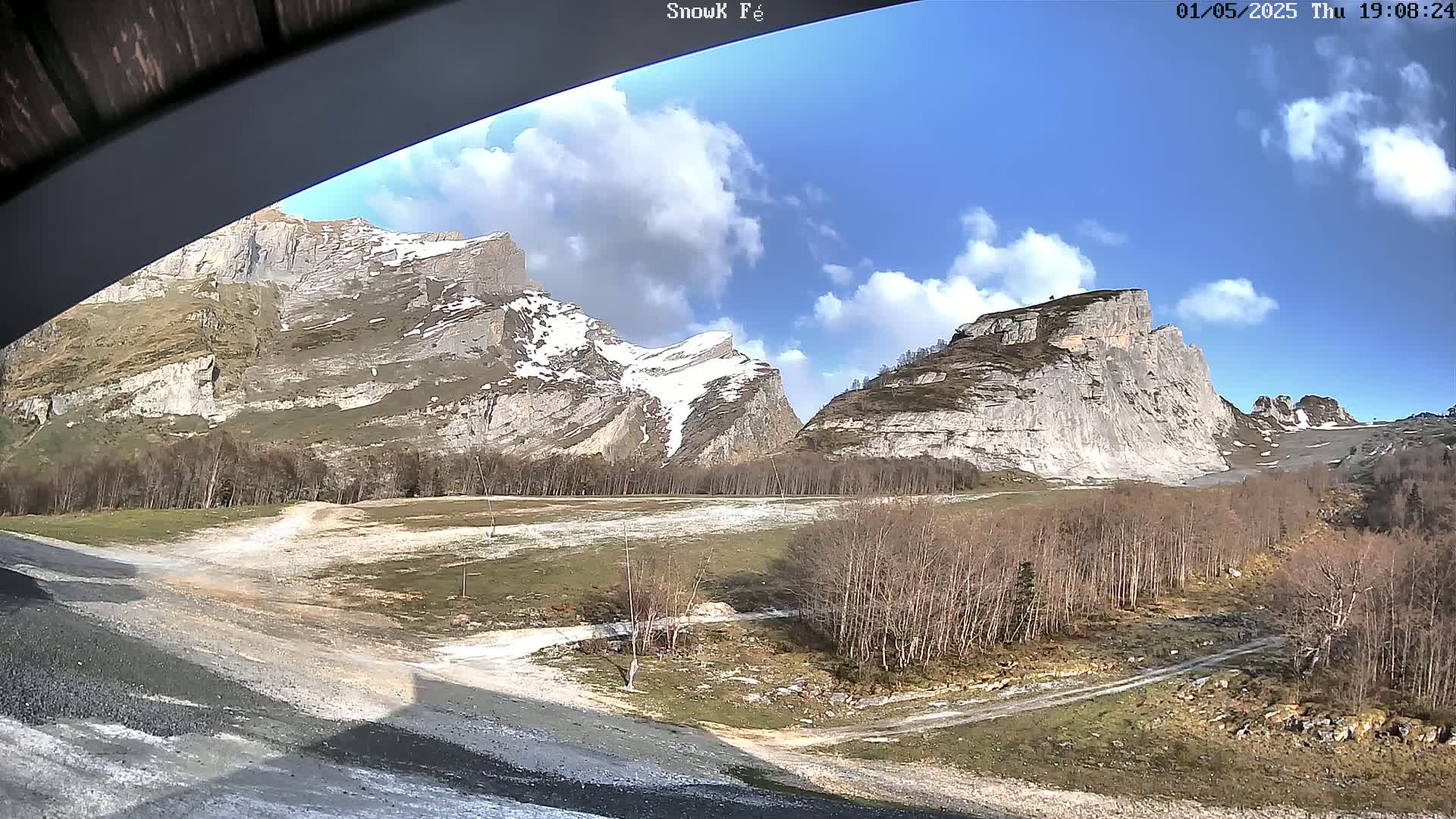 A mostly sunny day reveals snow-capped mountains and a valley with leafless trees and a dirt road.