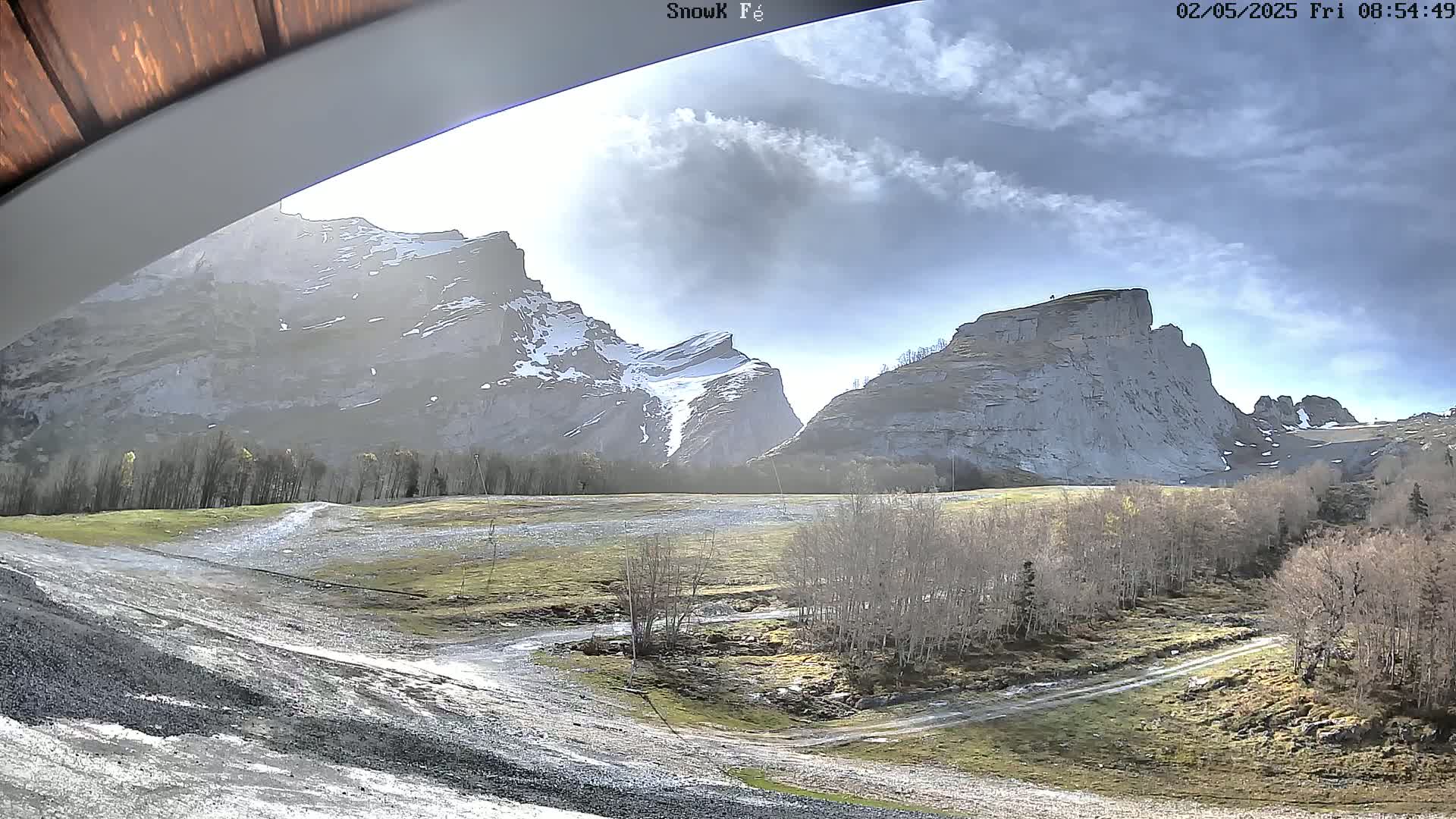 A partly cloudy sky overlooks a mountainous landscape with patches of snow, barren trees, and a gravel path.