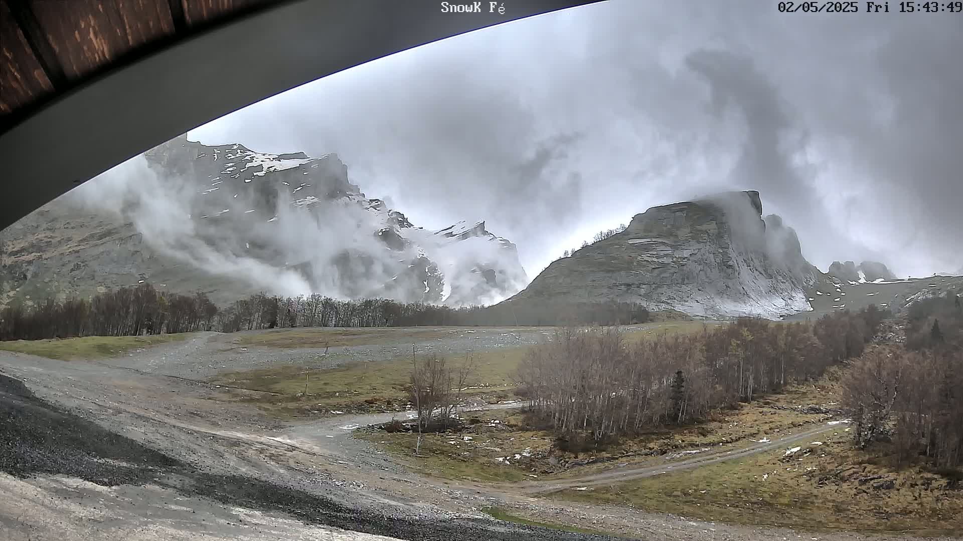 A cloudy, overcast day reveals snow-capped mountains partially obscured by fog and mist, with a valley below featuring sparse vegetation and a dirt road.