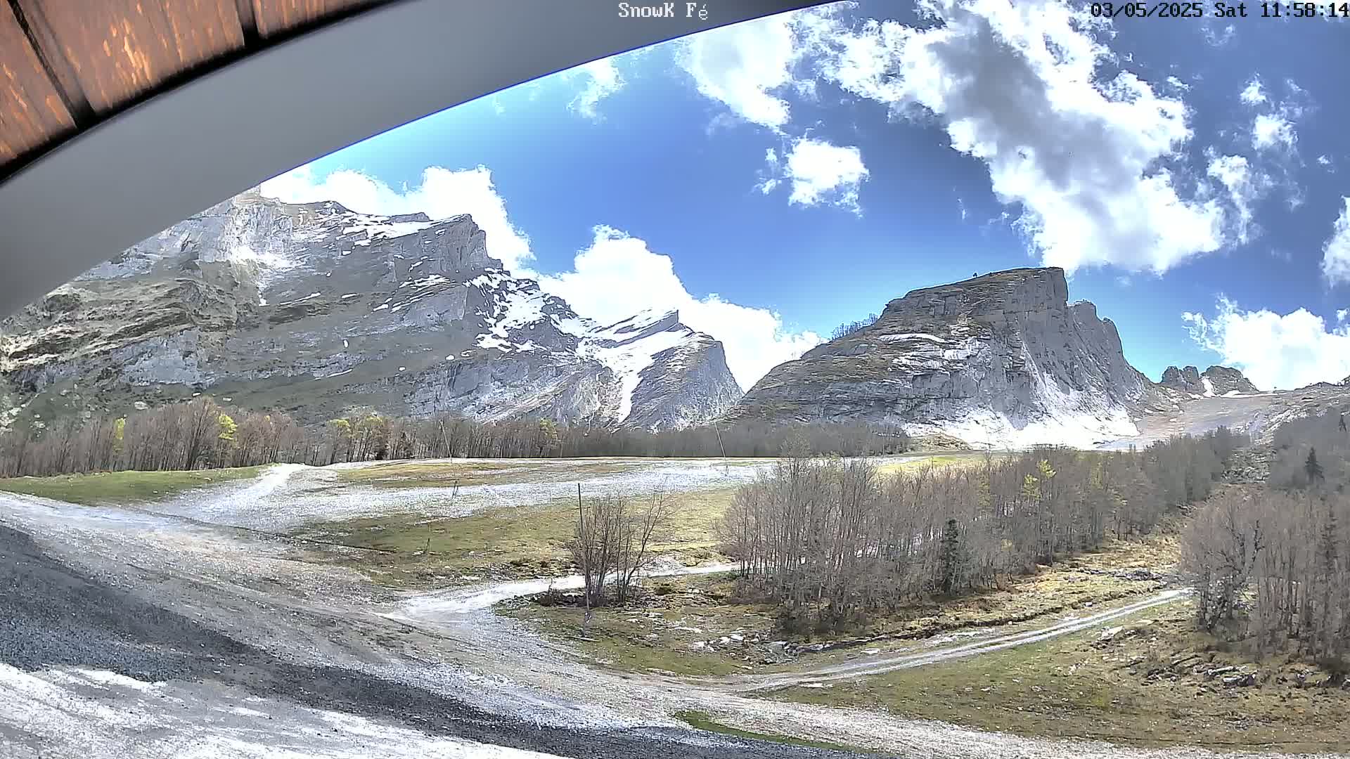 A partly cloudy sky overlooks a valley with barren trees, rocky mountains, and patches of snow.