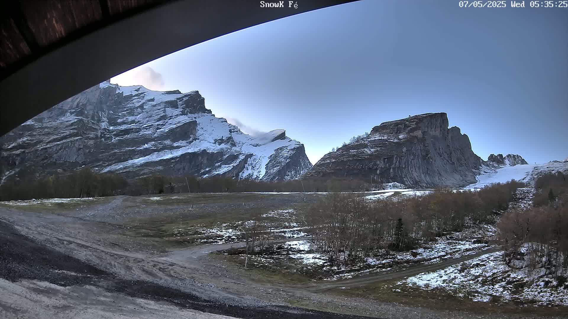 Snow-dusted mountains rise above a flat, partly snow-covered landscape with sparse trees and a dirt road under a clear, pale blue sky.