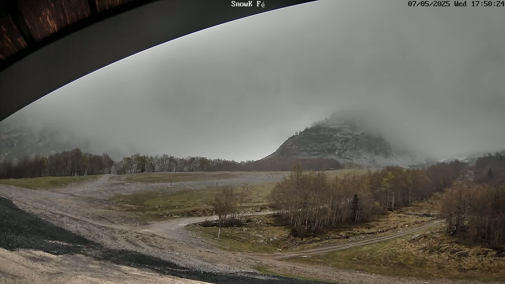 A foggy, overcast scene shows a rocky hill partially obscured by fog, with a dirt road and sparse trees in the foreground.