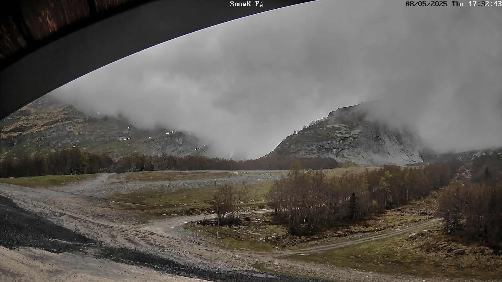 A foggy, overcast day reveals a rocky, mountainous landscape with sparse, leafless trees and a dirt road winding through a flat, grassy area in the foreground.