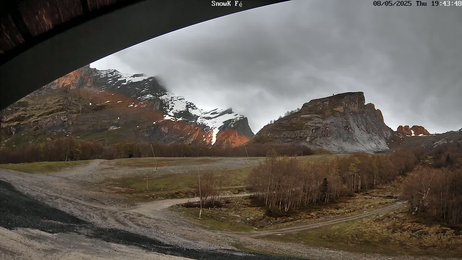 A view from under a structure shows a dirt road leading to a grove of leafless trees in front of snow-dusted, rocky mountains under an overcast sky.