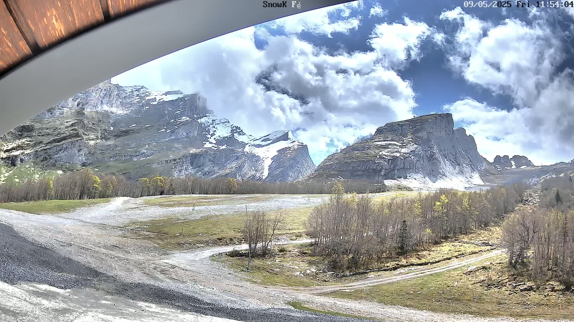 A partly cloudy sky overlooks a valley with snow-capped mountains, sparse trees, and a gravel road.