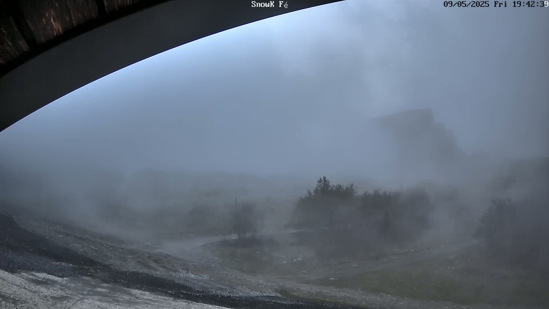 A view from under a curved structure shows a hazy, windy landscape with dark trees and a hill in the distance during a heavy storm.