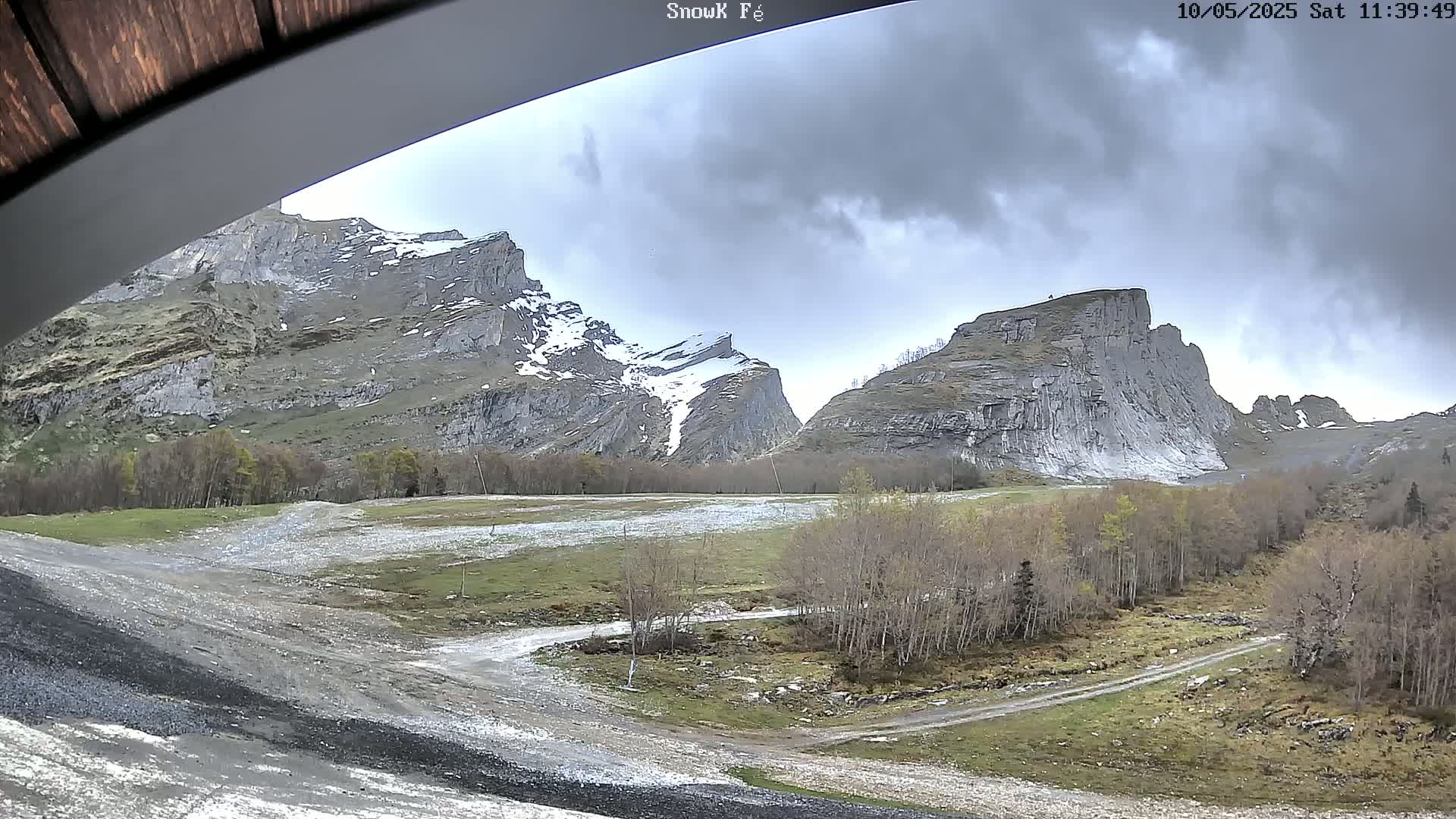 A view from under an overhang shows a valley with a dirt road winding through sparse trees and rocky, snow-dusted mountains under a cloudy sky.