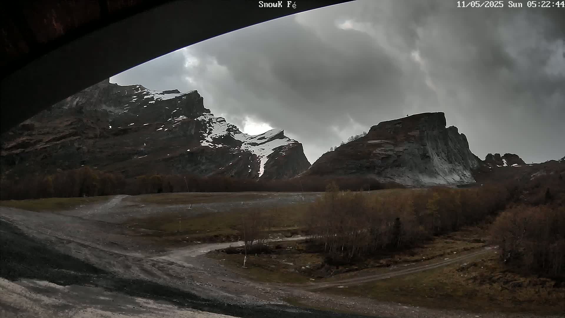 A view from under a structure shows a valley with a dirt road, sparse, leafless trees, and snow-capped mountains under an overcast sky.