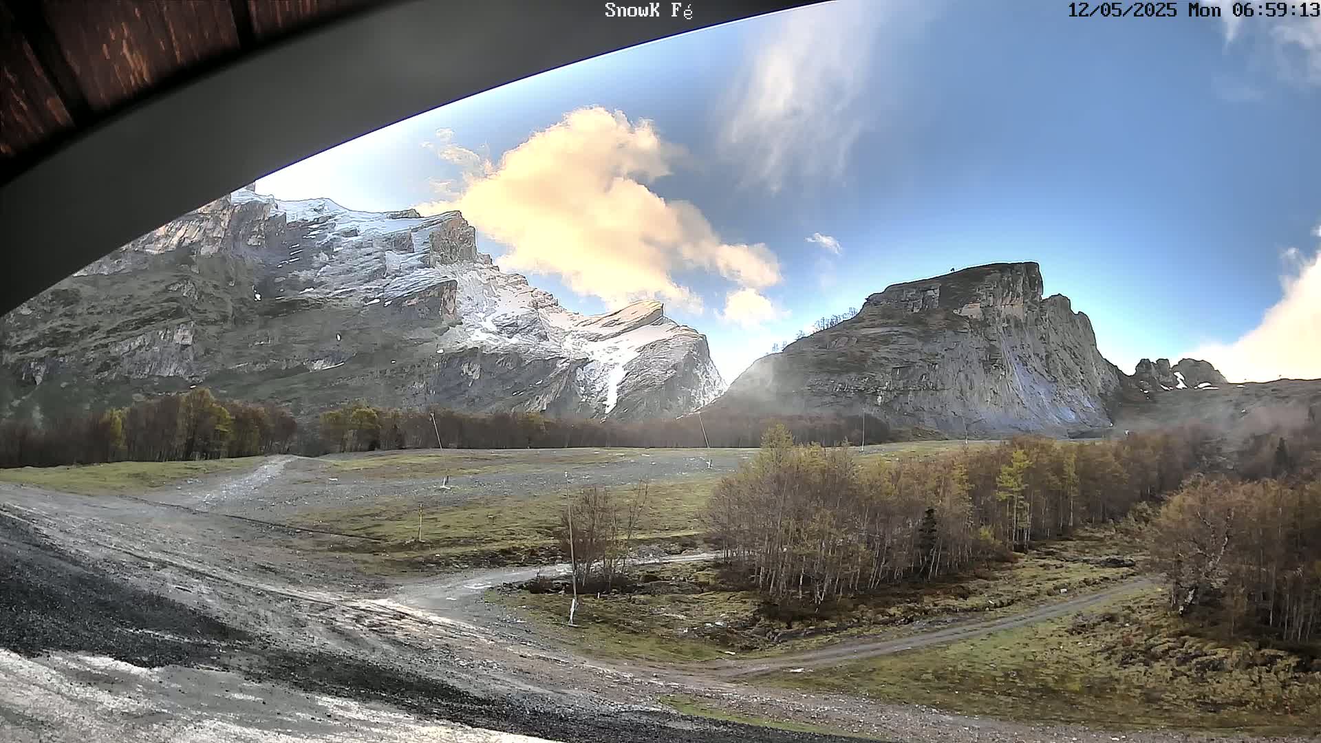 A partly cloudy day reveals snow-capped mountains and a valley with a dirt road and sparse, autumnal trees.