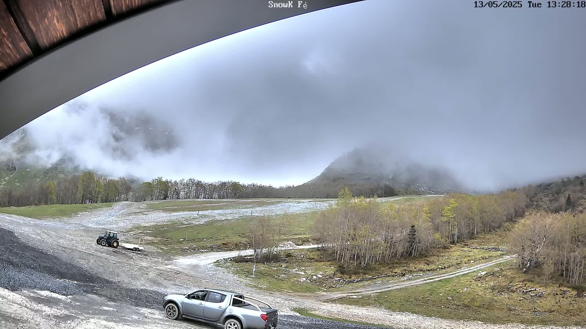 A gray pickup truck and a tractor with a trailer are parked on a gravel road leading to a mountain shrouded in fog.