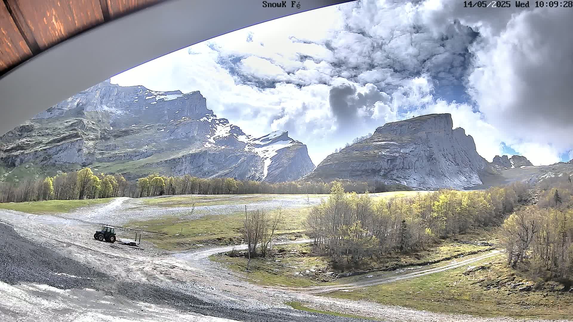 A tractor is parked on a gravel road in a valley, with snow-capped mountains and partly cloudy skies in the background.