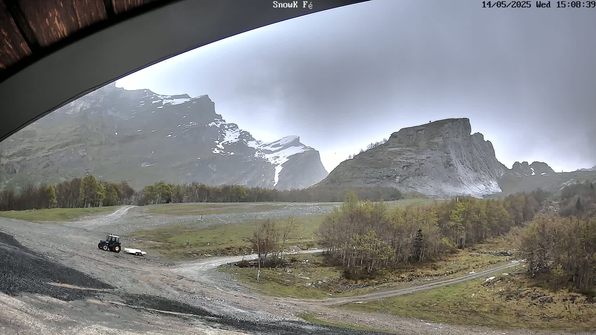 A tractor with a trailer is parked on a dirt road in a valley between snow-capped mountains on an overcast day.
