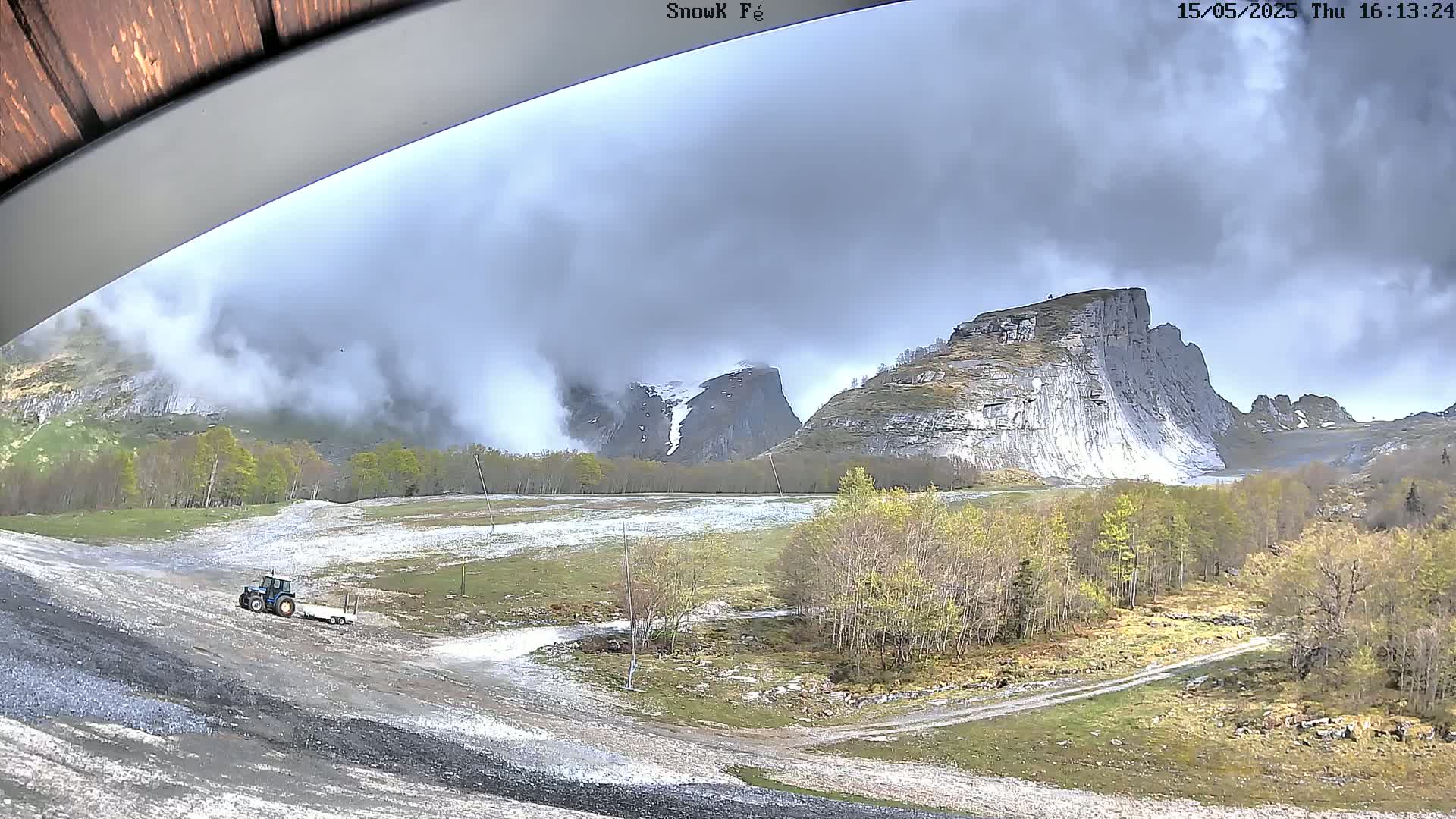 A tractor and trailer are visible on a mostly clear, rocky, and grassy area in front of a mountainous landscape partially covered in clouds.