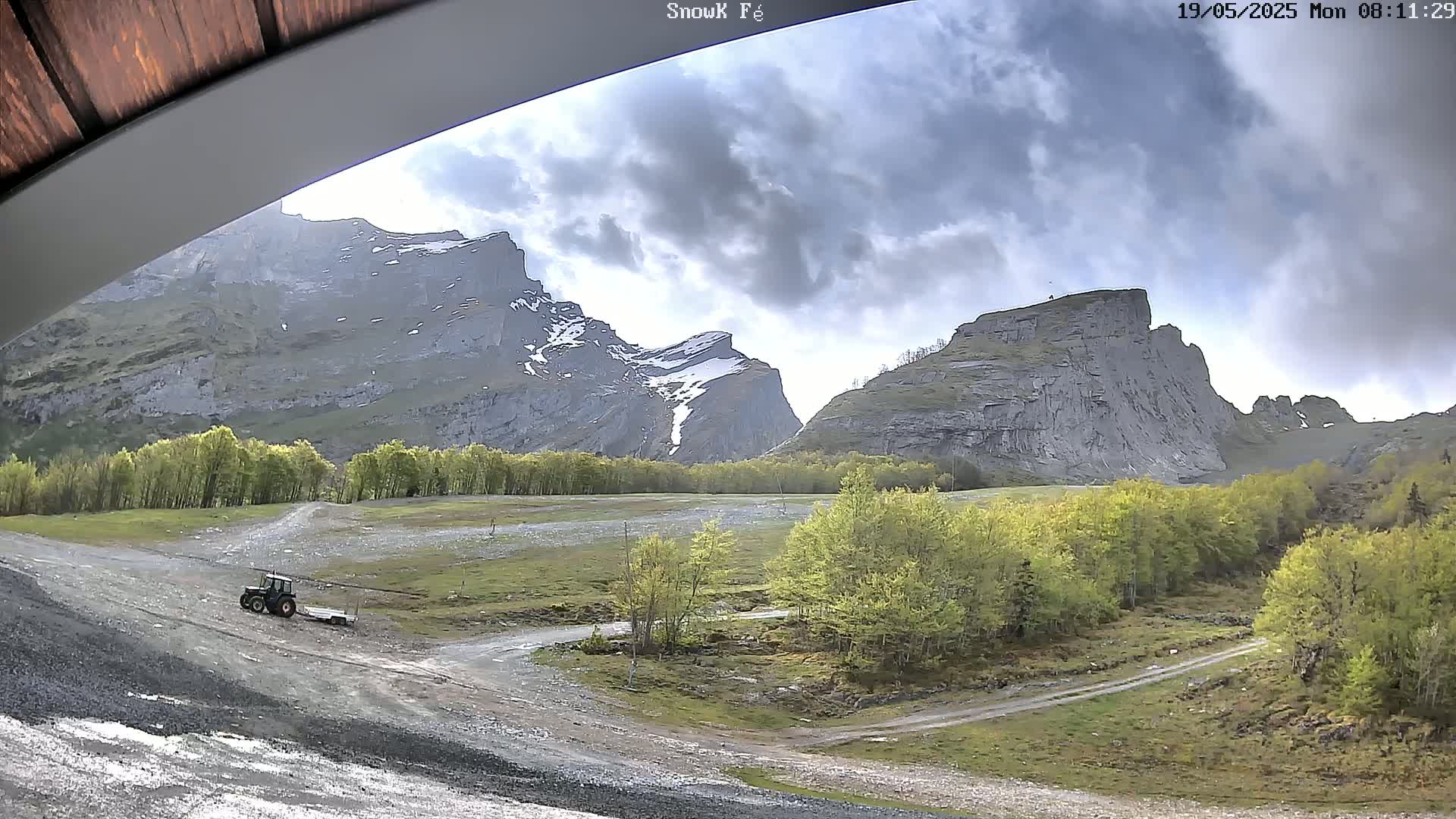 A tractor is parked on a gravel road amidst a valley of green trees and towering, gray, rocky mountains under a partly cloudy sky.