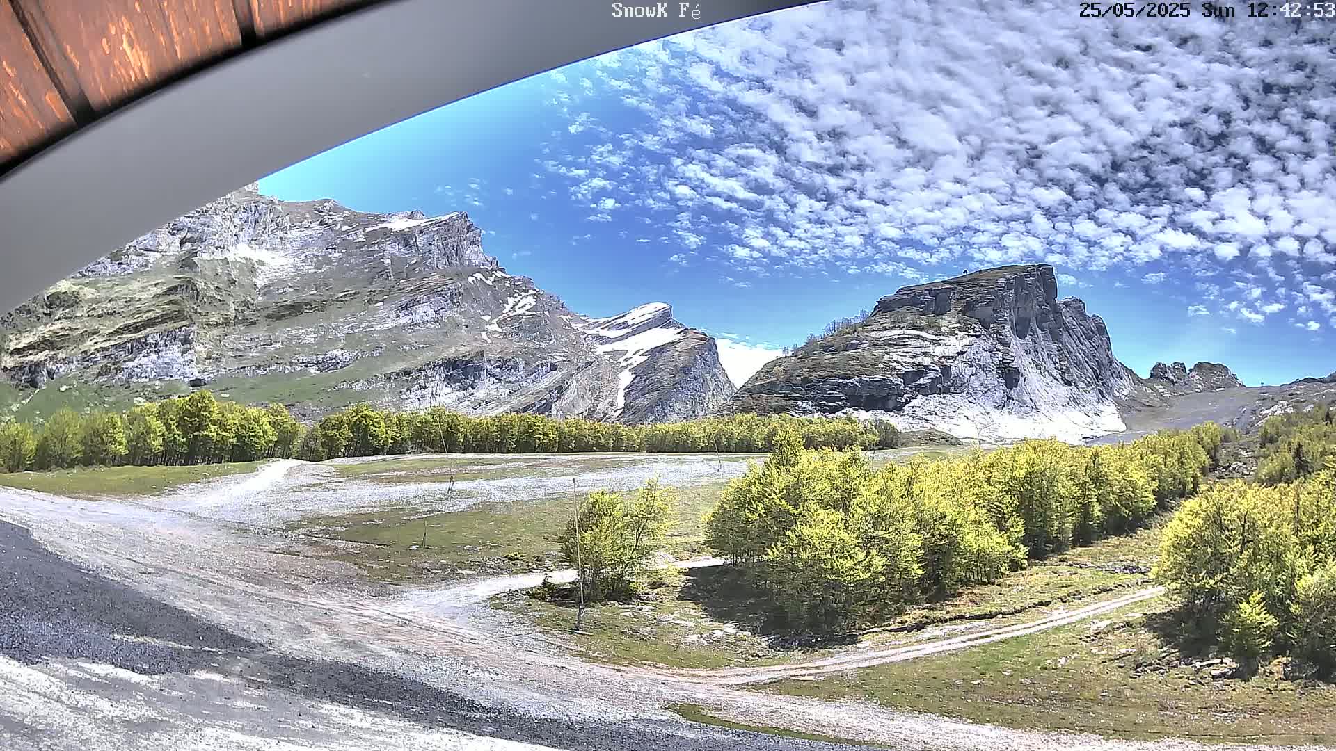 A sunny day reveals a valley with patches of grass and bright green trees nestled between large, rocky mountains.