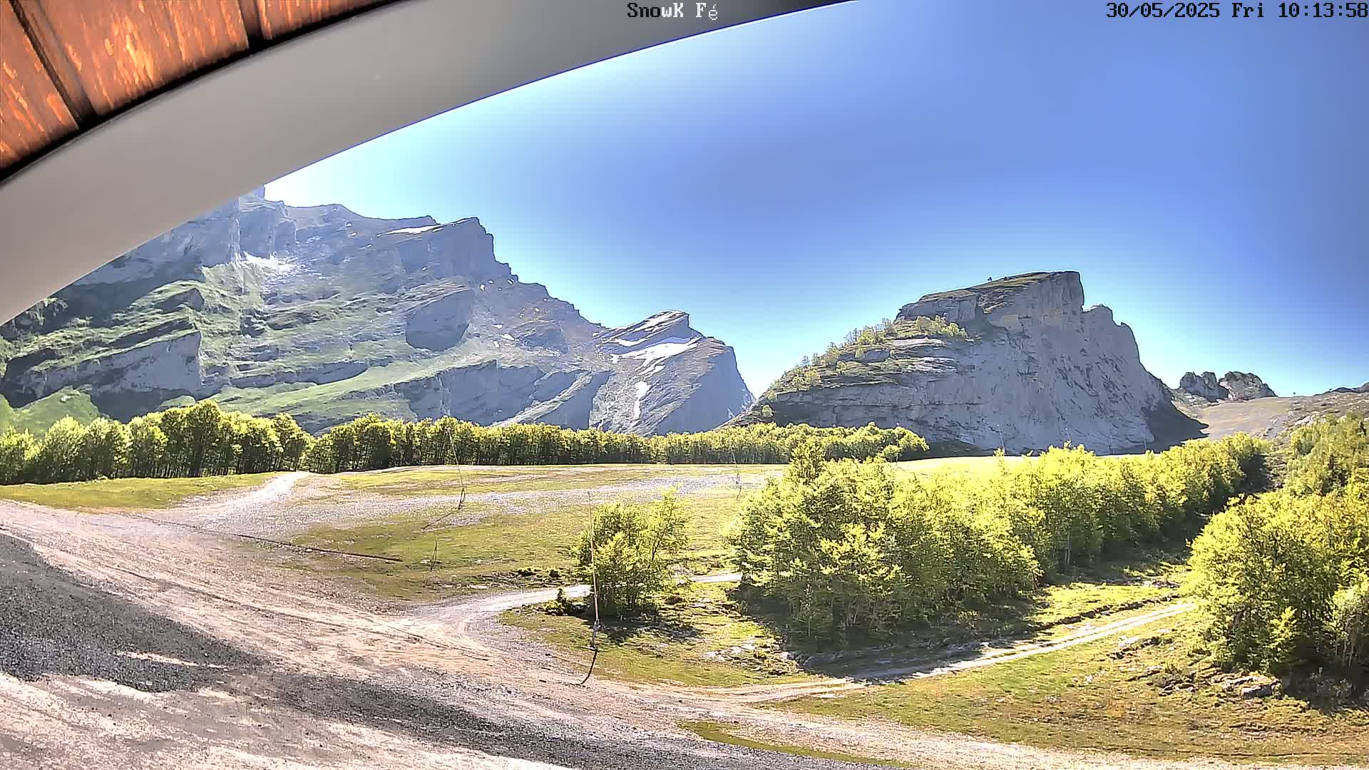A sunny view from under an awning shows a grassy valley with a dirt road, surrounded by green trees and towering, rocky mountains.