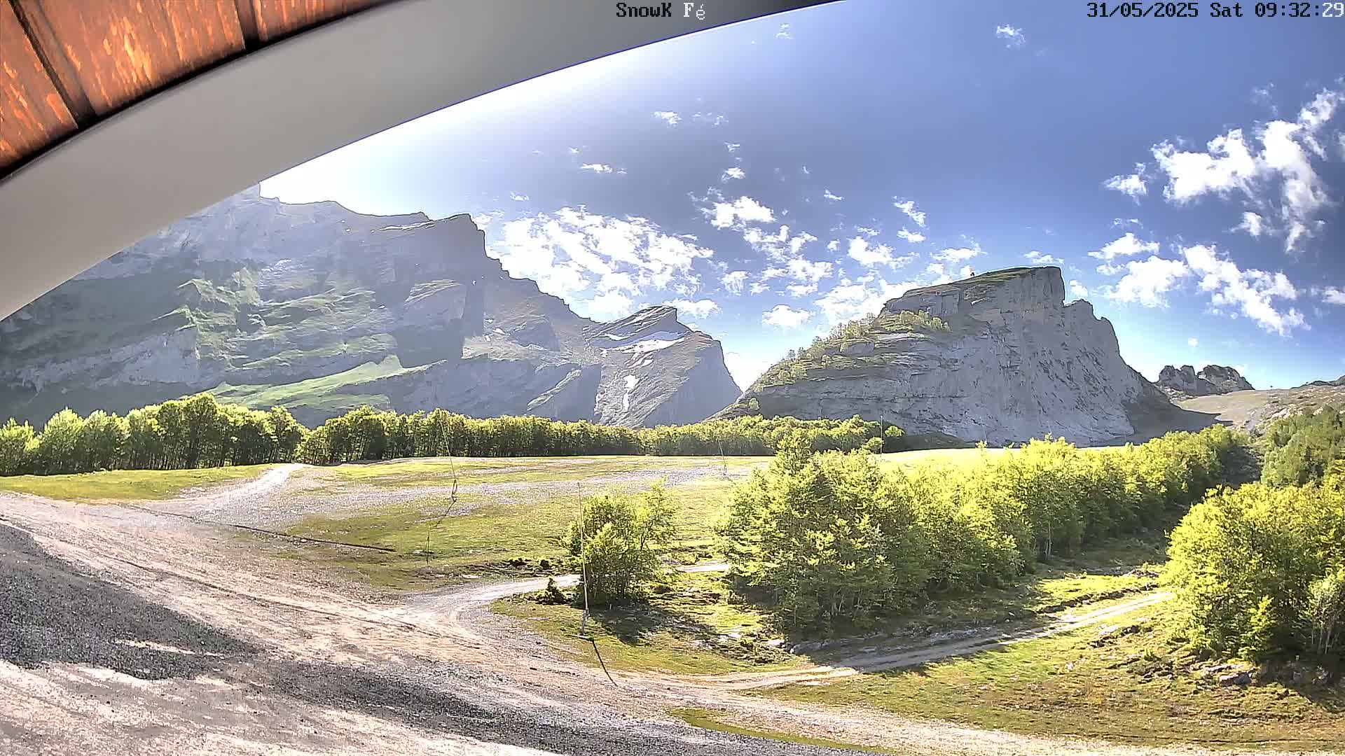 A sunny day reveals a mountain valley with a grassy field, dirt roads, and trees, backed by large, light-grey rocky mountains.