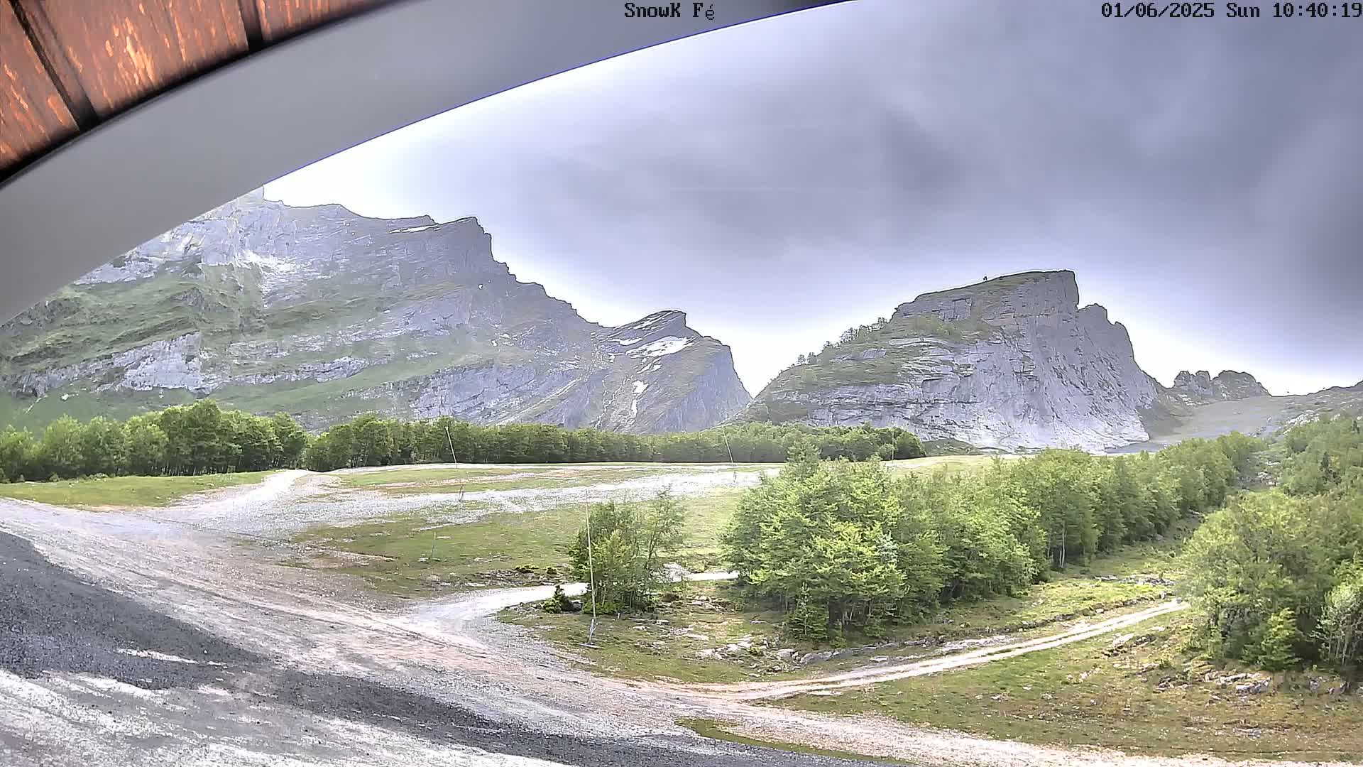 A view from under a roof shows a flat, grassy area with dirt roads between two large, gray, rocky mountains under an overcast sky.