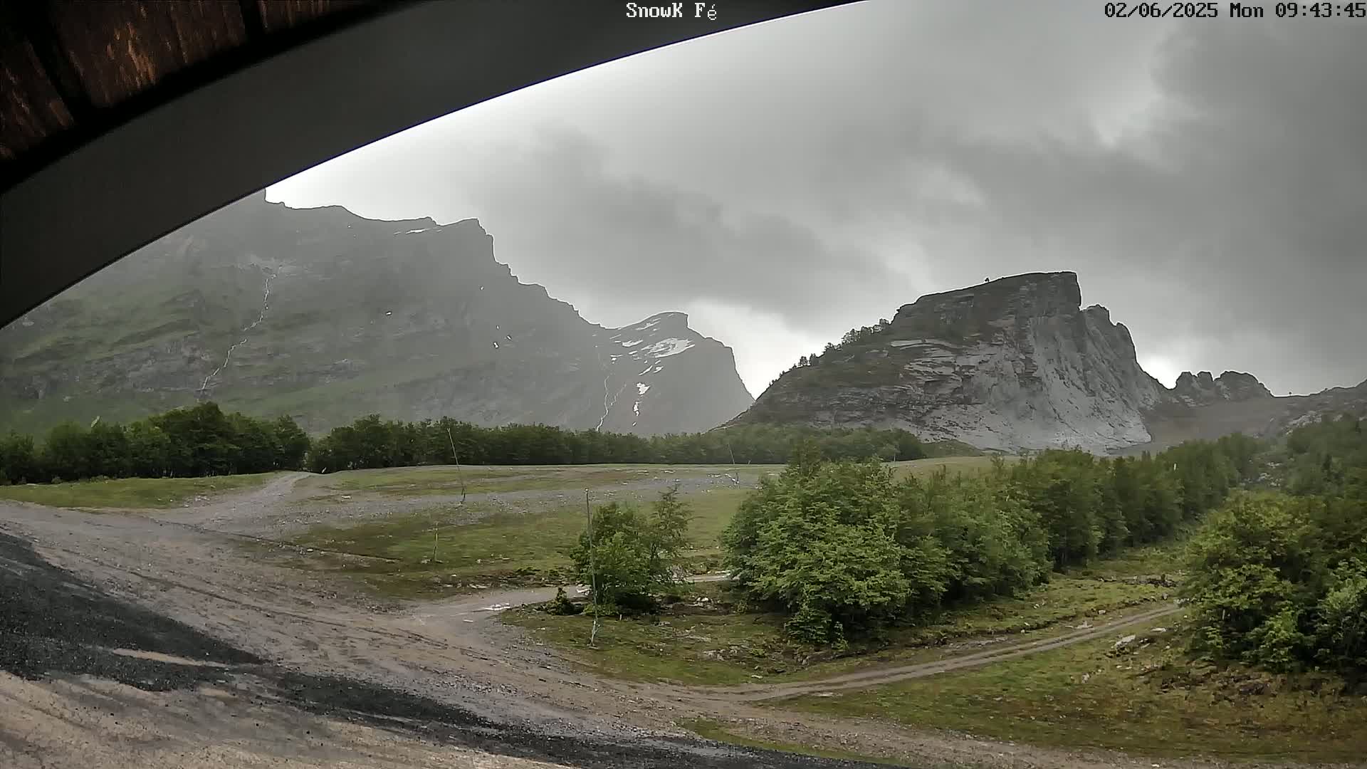 A view from under an overhang shows a valley with green trees and a dirt road, nestled between imposing, gray, rain-swept mountains.