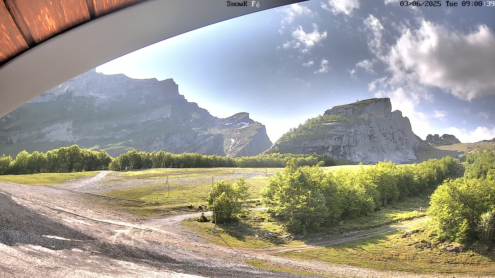 A sunny day reveals a rocky mountain landscape with green trees and a dirt road winding through the foreground.