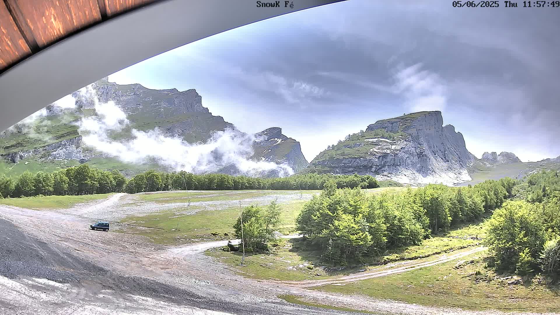 A dark-colored SUV is parked on a gravel road in a mountainous valley, with partly cloudy skies and low-lying clouds partially obscuring the rocky peaks.