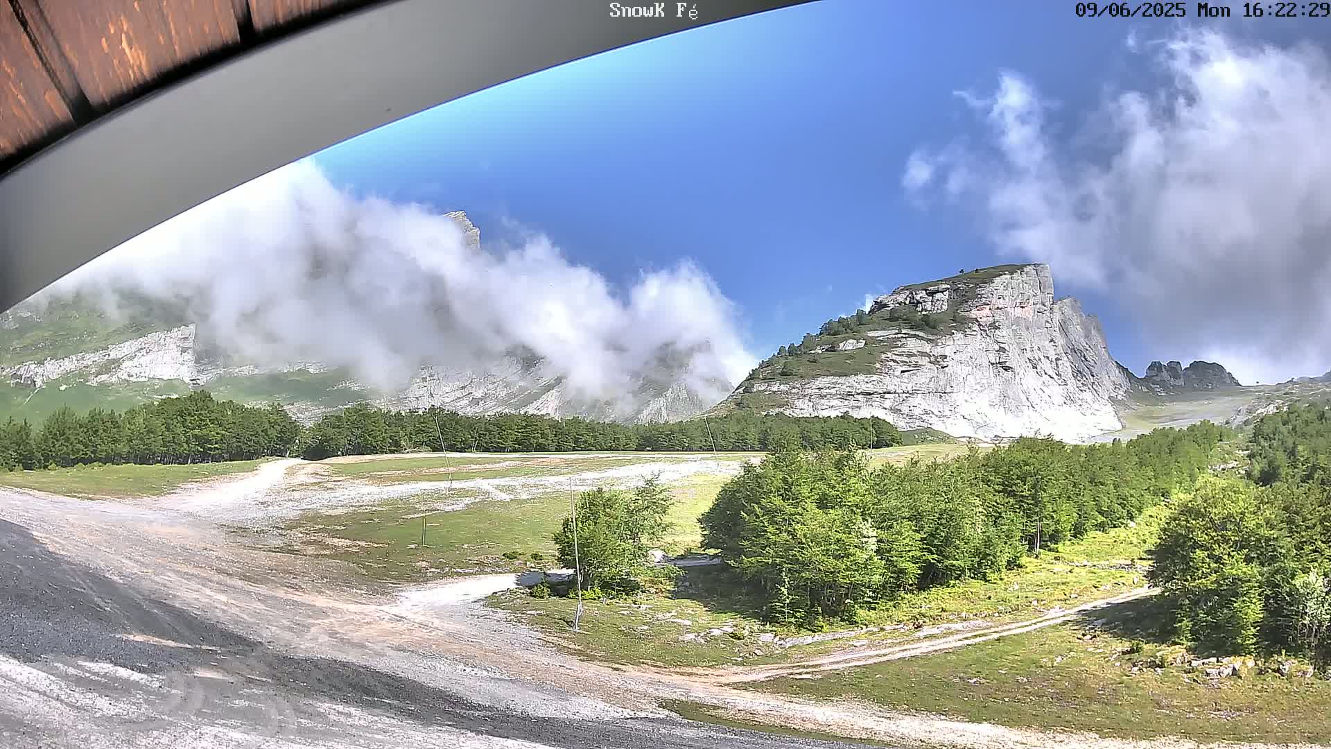 A partly cloudy day reveals a mountainous landscape with a dirt road winding through a green forest toward a prominent, light-colored cliff face.