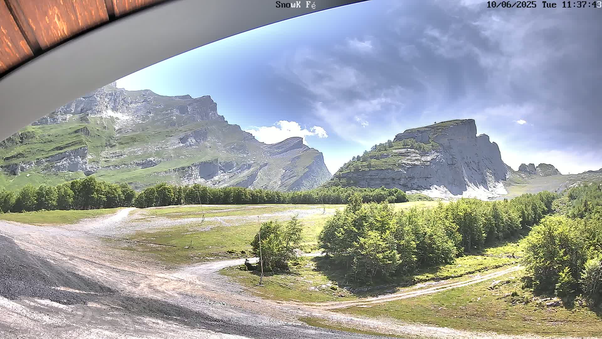 A partly cloudy day reveals a mountain valley with a gravel road and green trees nestled between towering, light gray rock formations.