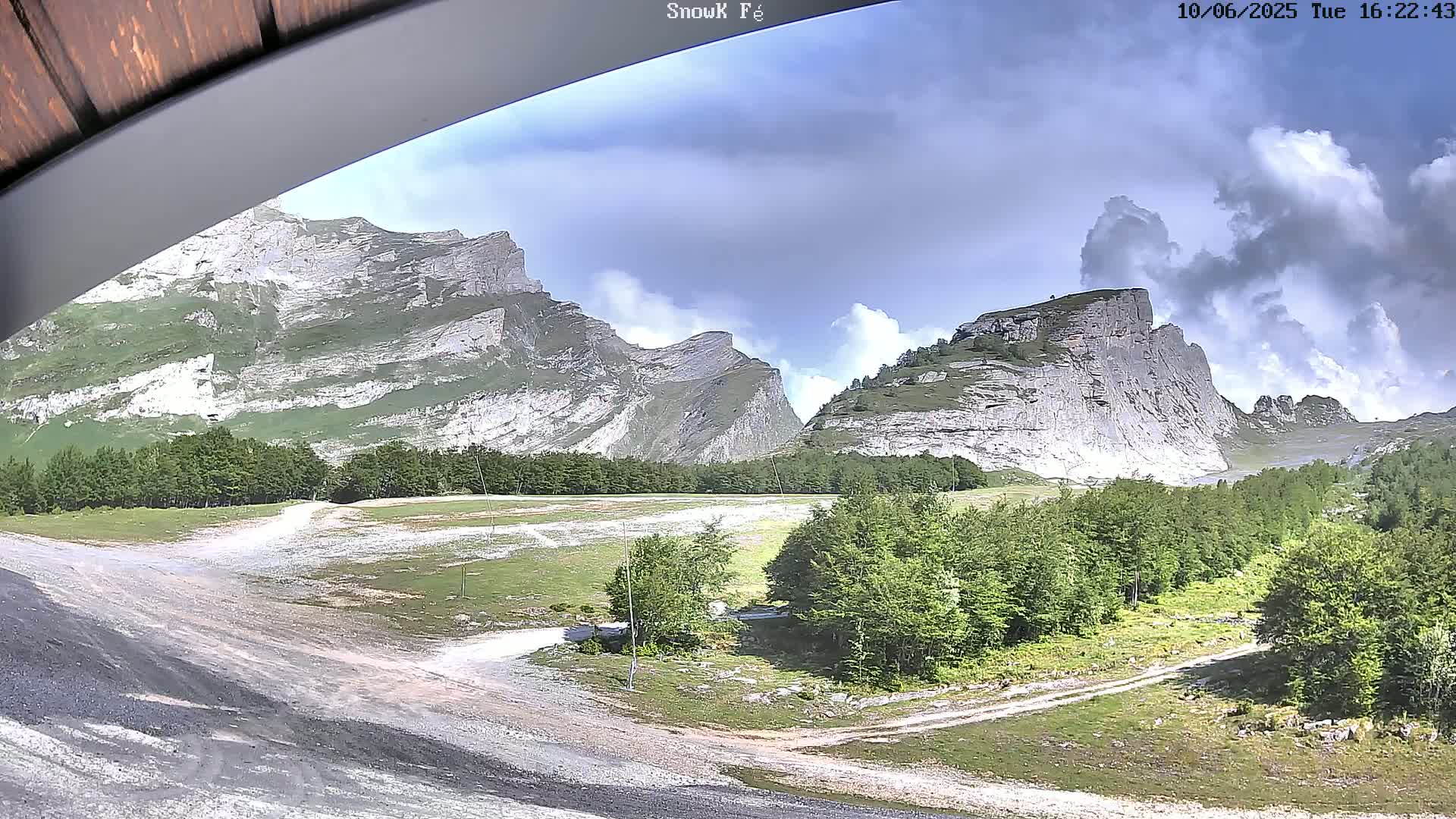 A partly cloudy sky overlooks a mountain valley with rocky peaks, green trees, and a dirt road.