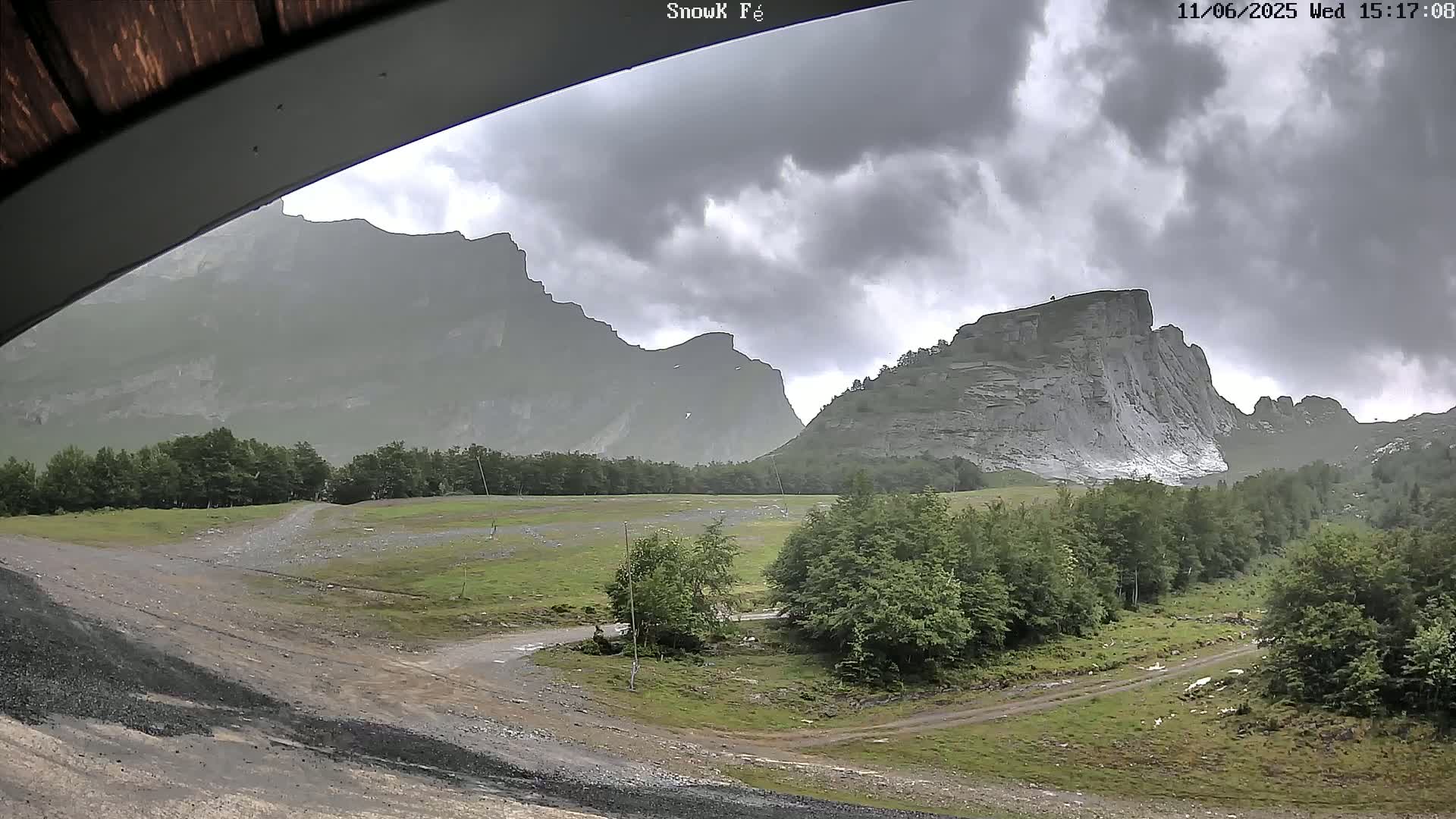 A view from under an overhang shows a dirt road and grassy area leading toward imposing, gray, rocky mountains under a cloudy sky.