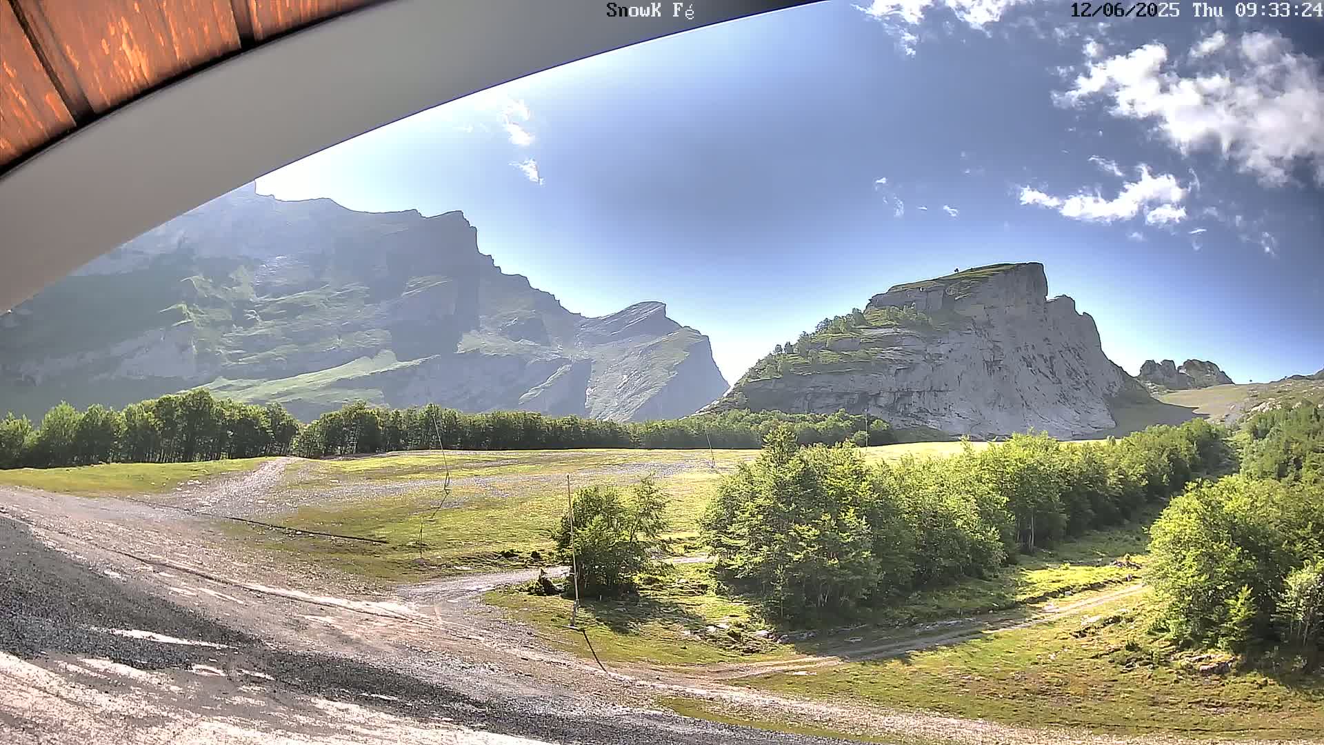 A sunny day reveals a valley with green vegetation, a dirt road, and towering, light-grey rocky mountains in the background.