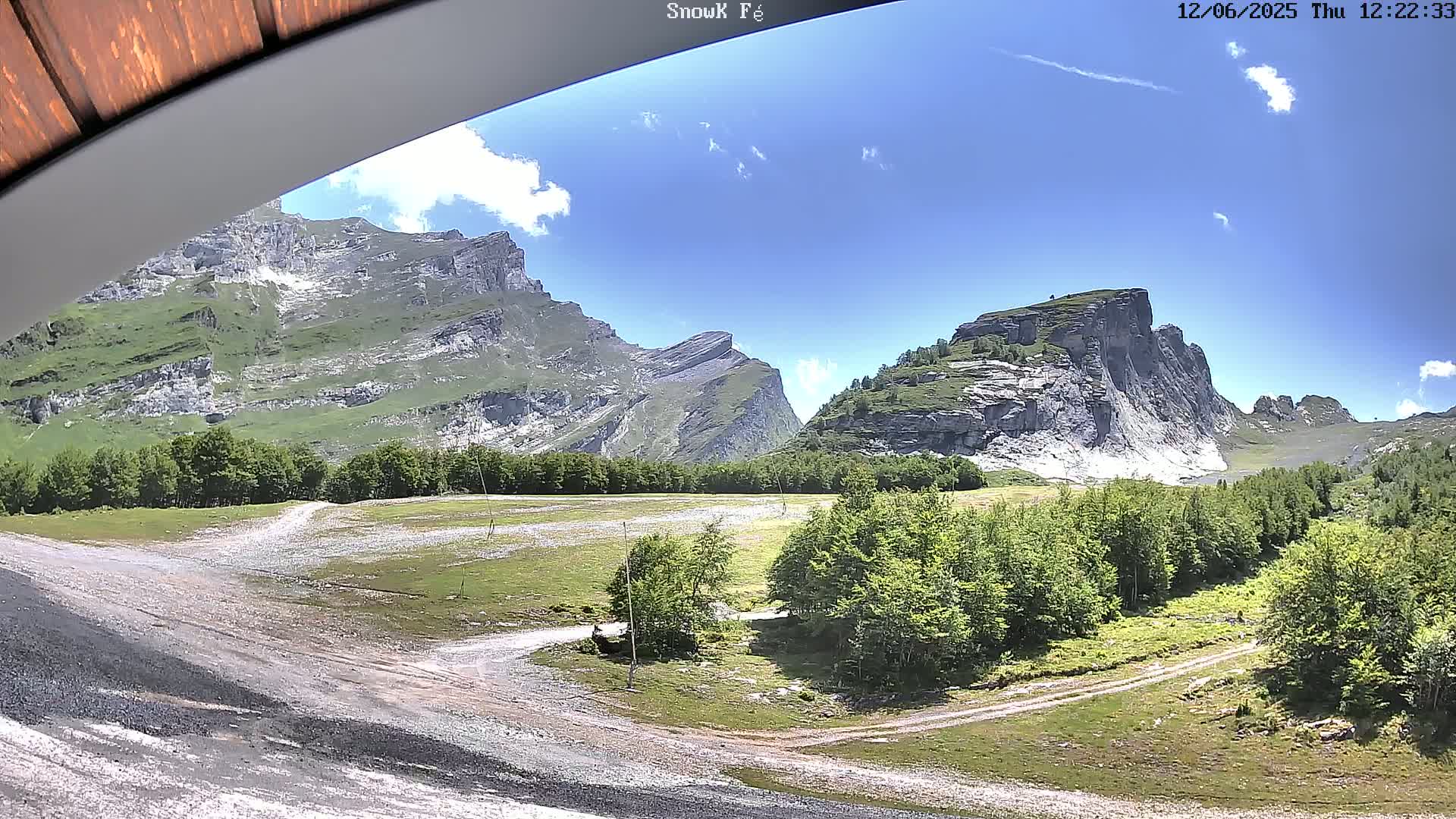 A sunny day reveals a mountain valley with a dirt road winding through green fields and forests, framed by towering, rocky mountains.
