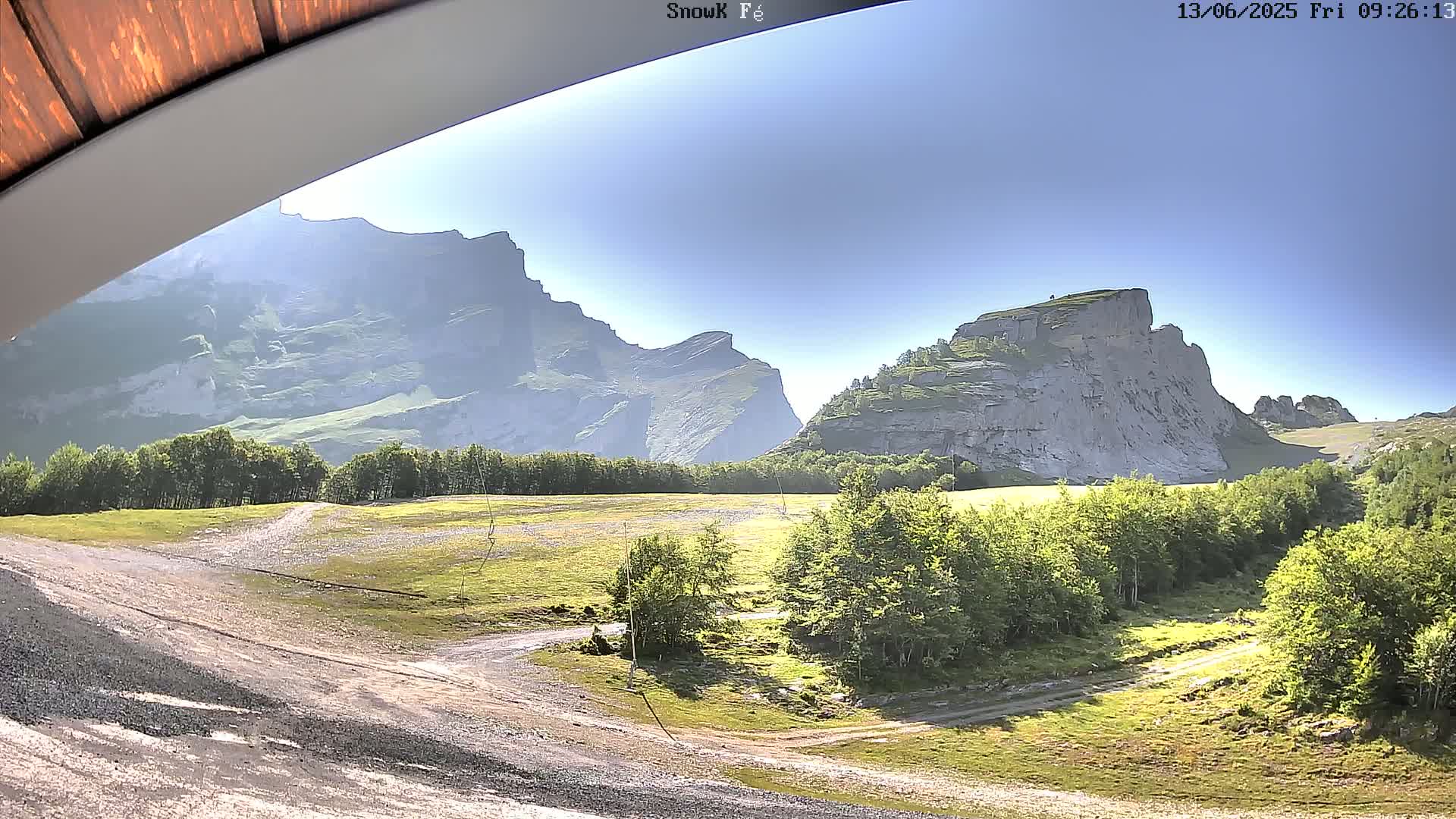 A sunny view from under a wooden overhang shows a grassy valley with dirt roads and green trees, and large, light-grey rocky mountains in the background.