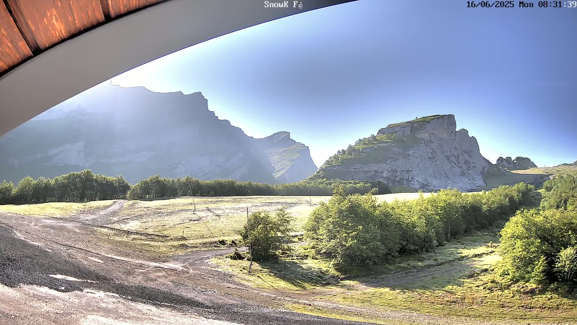 A sunny view from under a roof shows a flat, grassy area with a dirt road, bordered by green trees and large, light-grey mountains in the background.