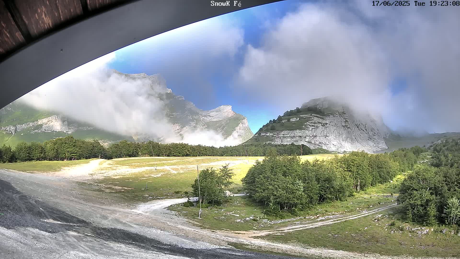 A partly cloudy day reveals mountain peaks partially obscured by clouds and mist, with a valley below containing green trees and dirt roads.