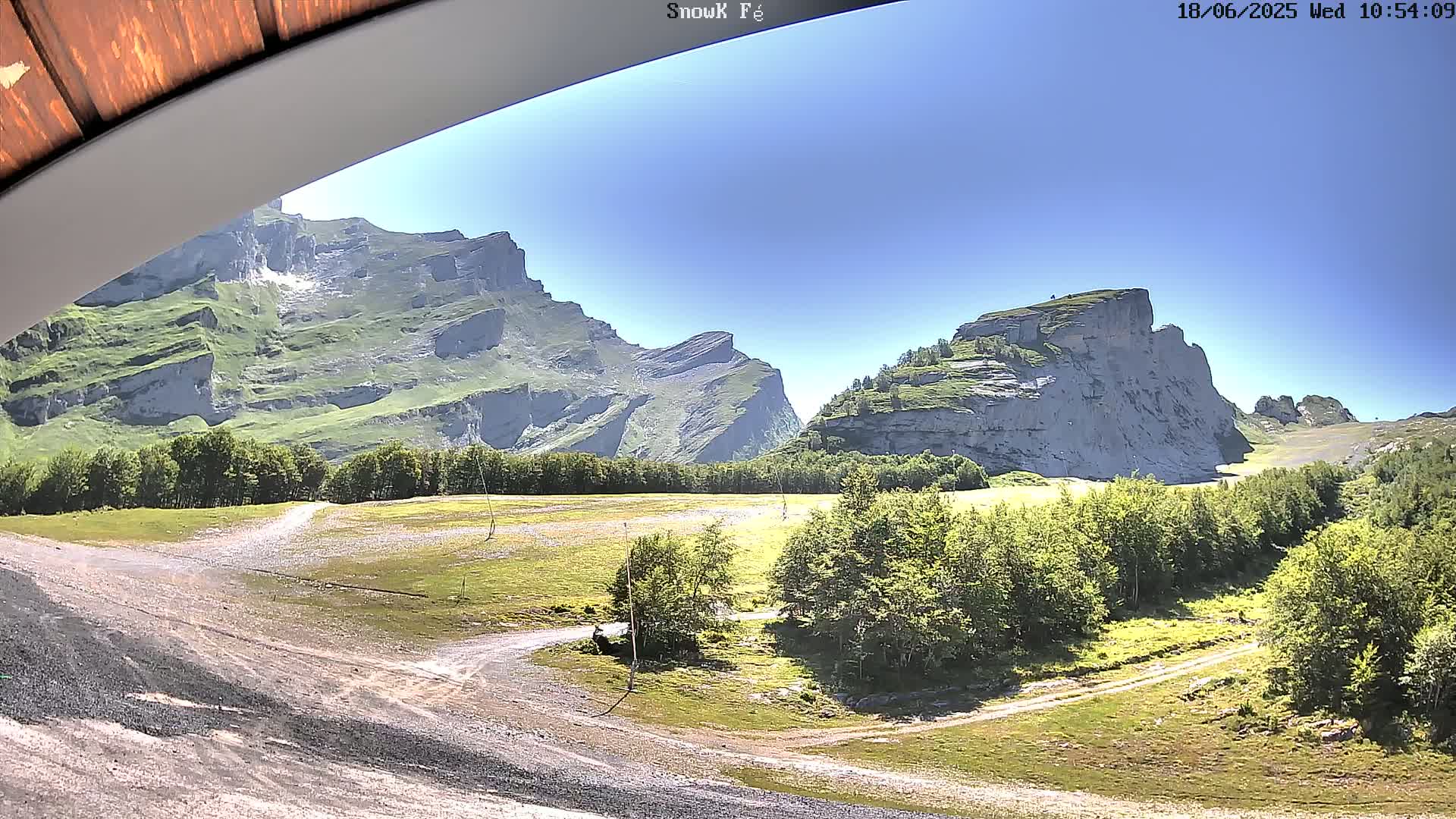 A sunny view of a mountainous landscape with green vegetation and dirt roads, seen from under a wooden overhang.