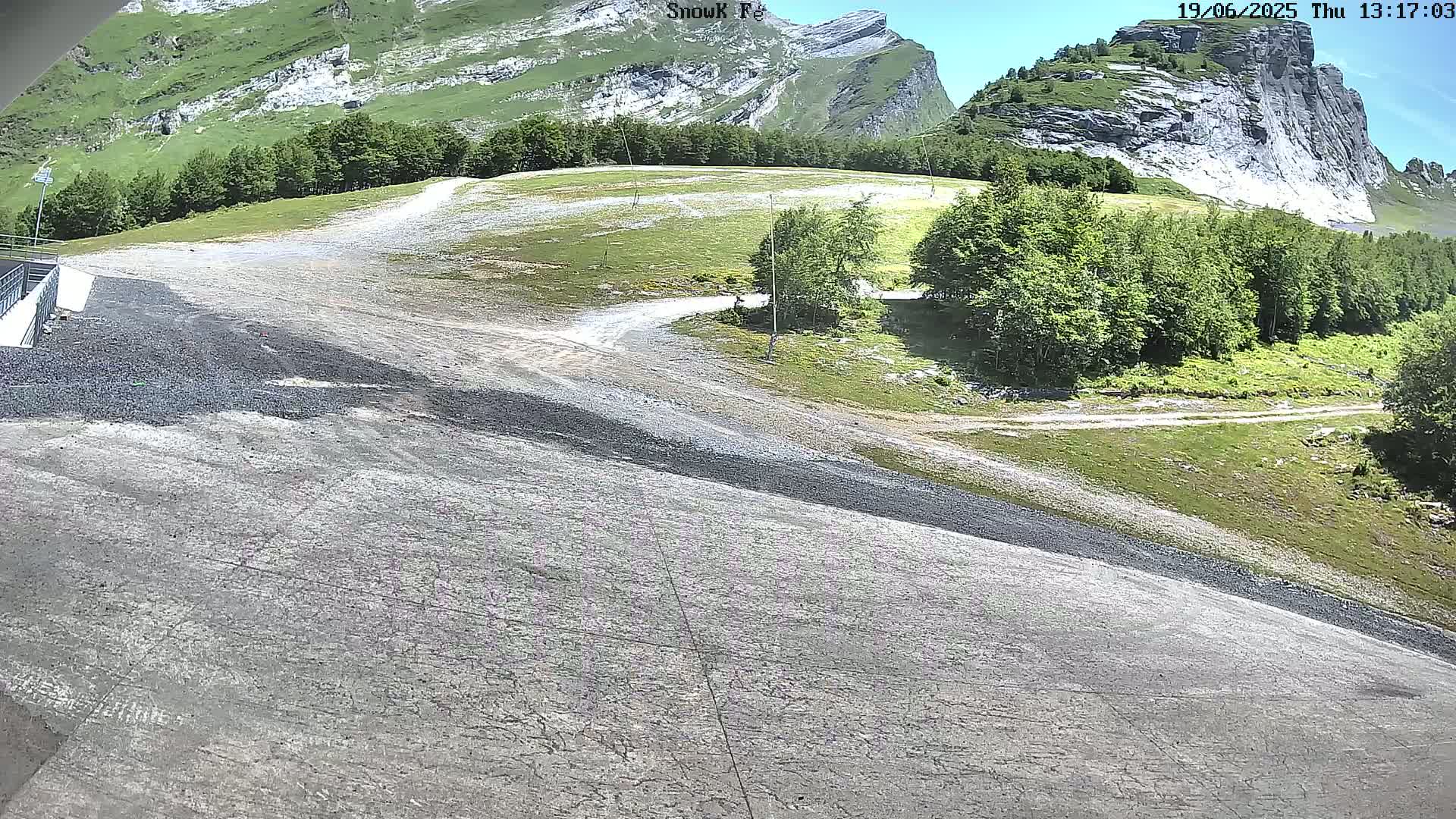 A rocky, gravelly slope descends toward the camera, revealing a verdant valley with a rocky mountain in the background under a clear sky.