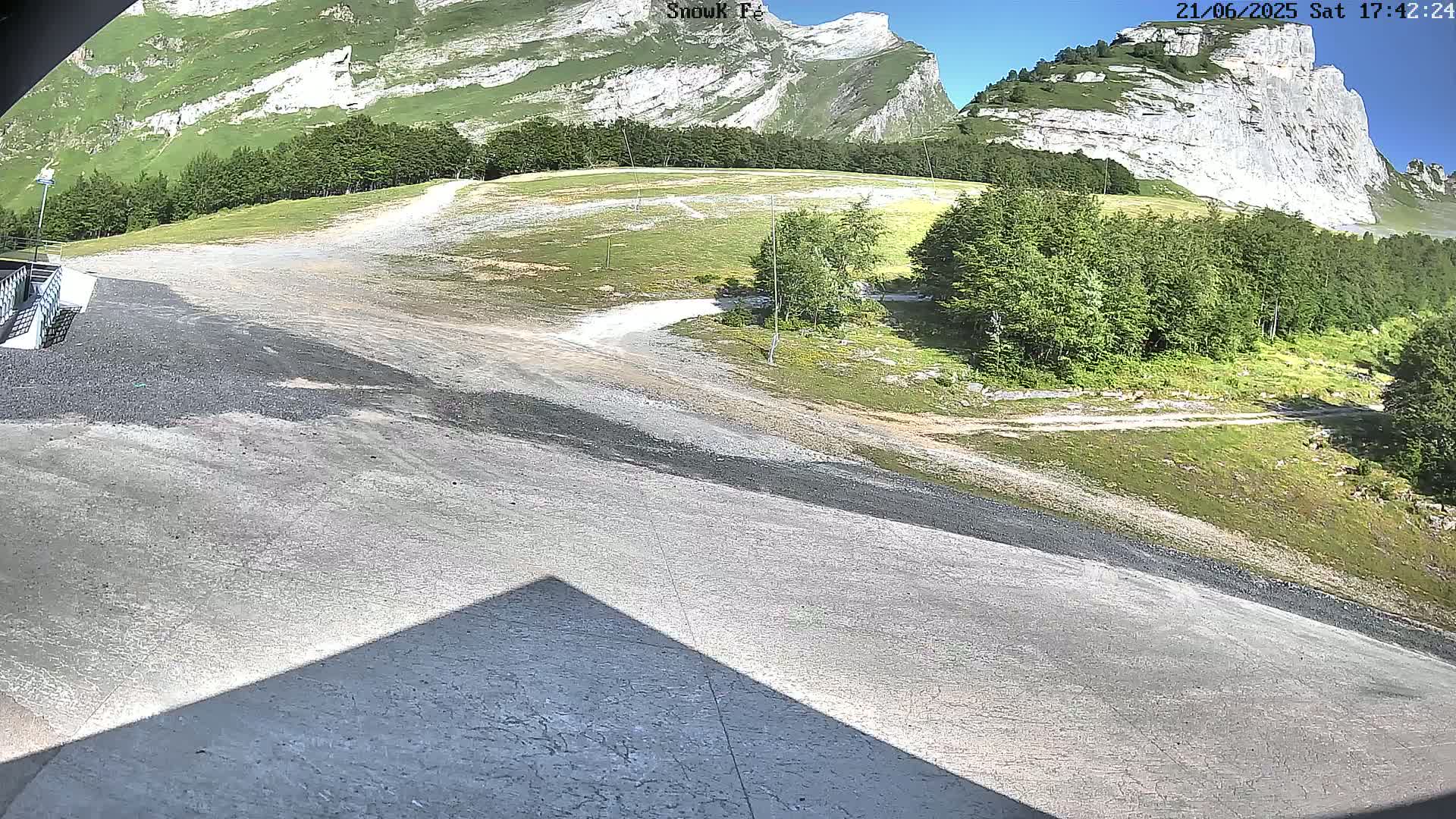 A paved area slopes downward toward a grassy expanse, with a backdrop of rocky mountains under a sunny sky.