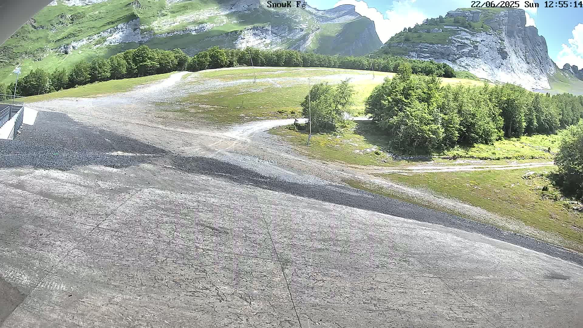 A mostly sunny outdoor scene shows a gravel slope leading to a grassy area with trees and rocky mountains in the background.