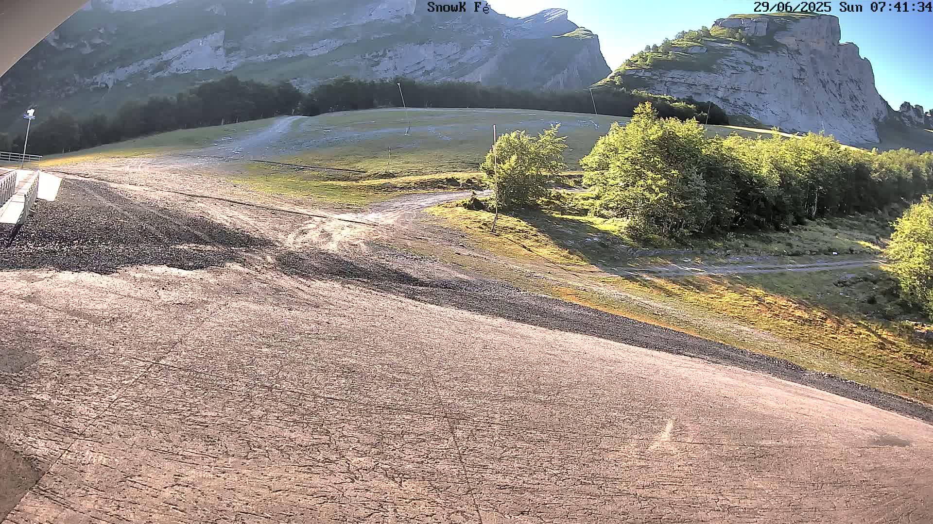 A mostly sunny, high-angle view of a gravelly, sloped path leading towards a grassy area with trees and large, rocky mountains in the background.