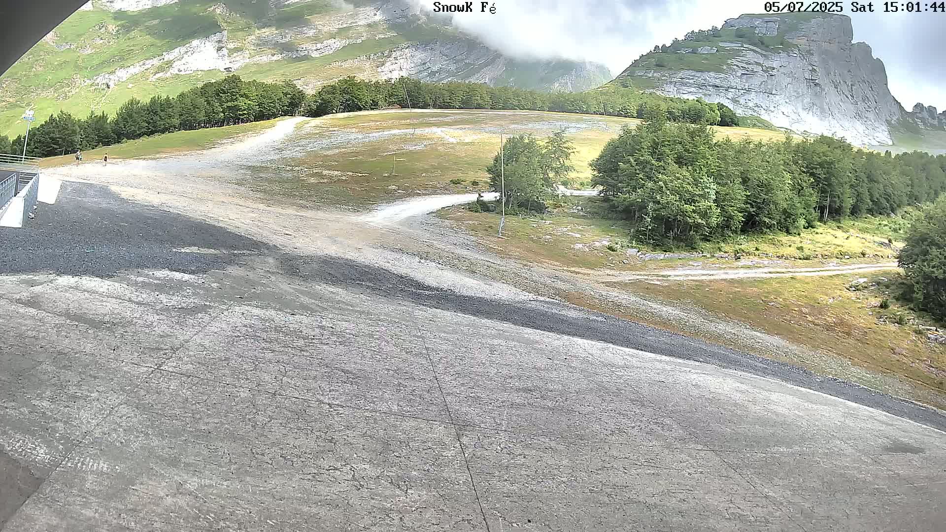 A paved area slopes downward toward a gravel path winding through a grassy field and a forest, with a large, light-colored rock formation and cloudy skies in the background.