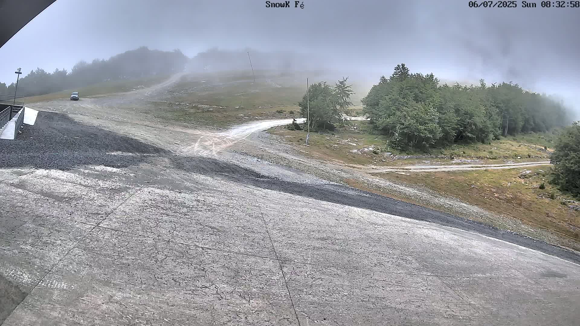 A foggy, overcast day reveals a gravel road winding through a mountainous area with sparse vegetation and a paved area in the foreground.