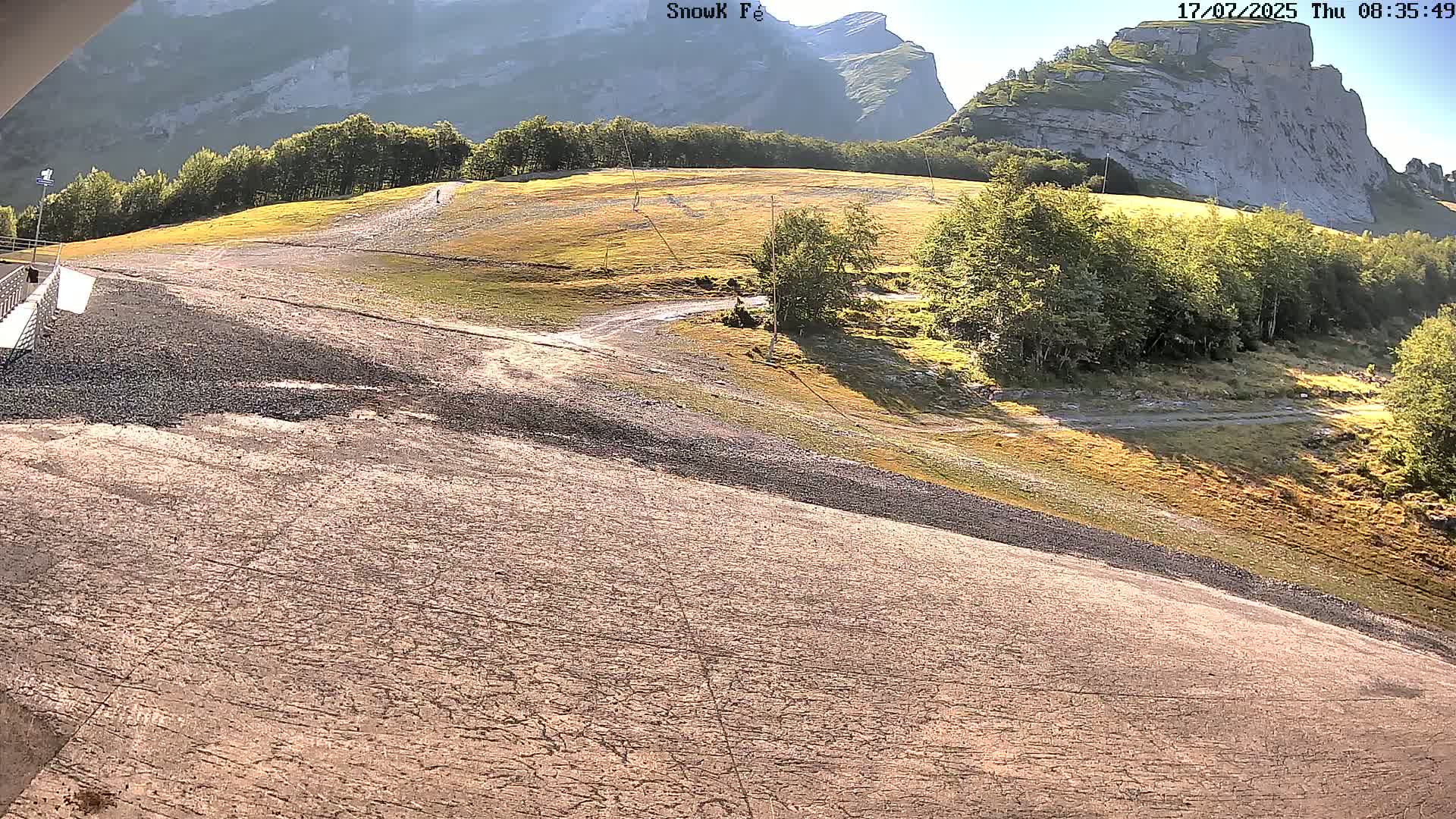 A dry, gravelly slope descends toward a viewer, leading to a grassy area with trees and rocky mountains in the background under a sunny sky.